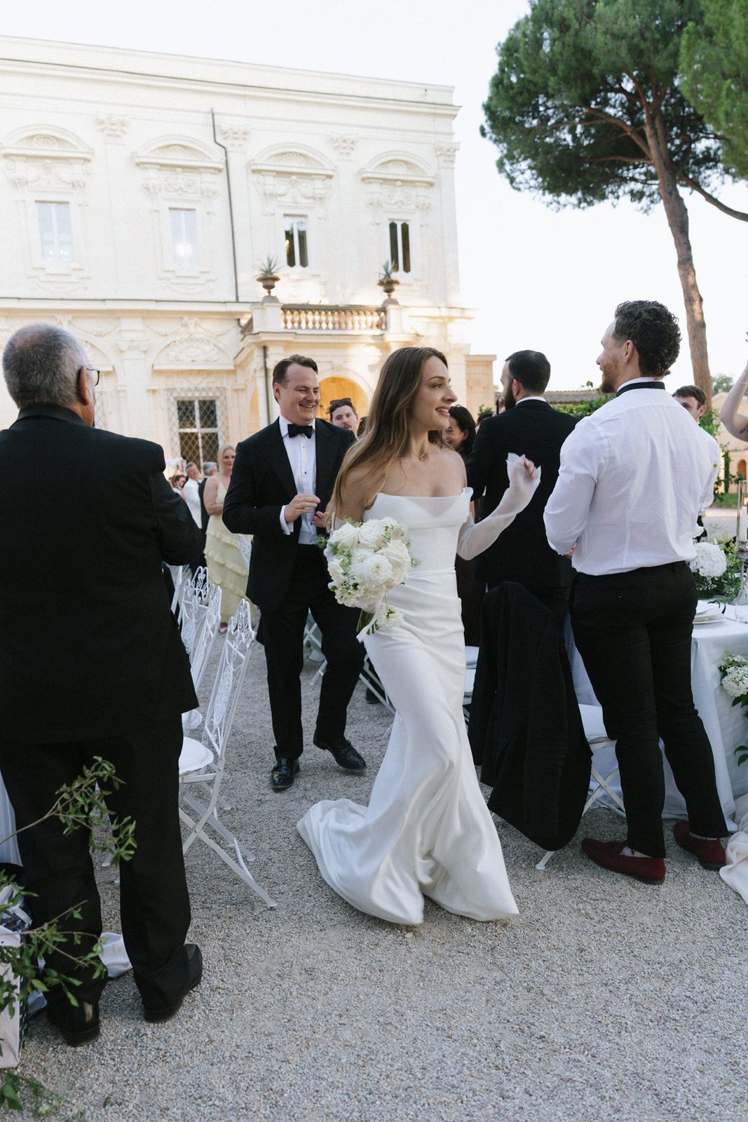 Bride in white off-shoulder gown walks with groom in tuxedo at elegant outdoor wedding venue in Italy.