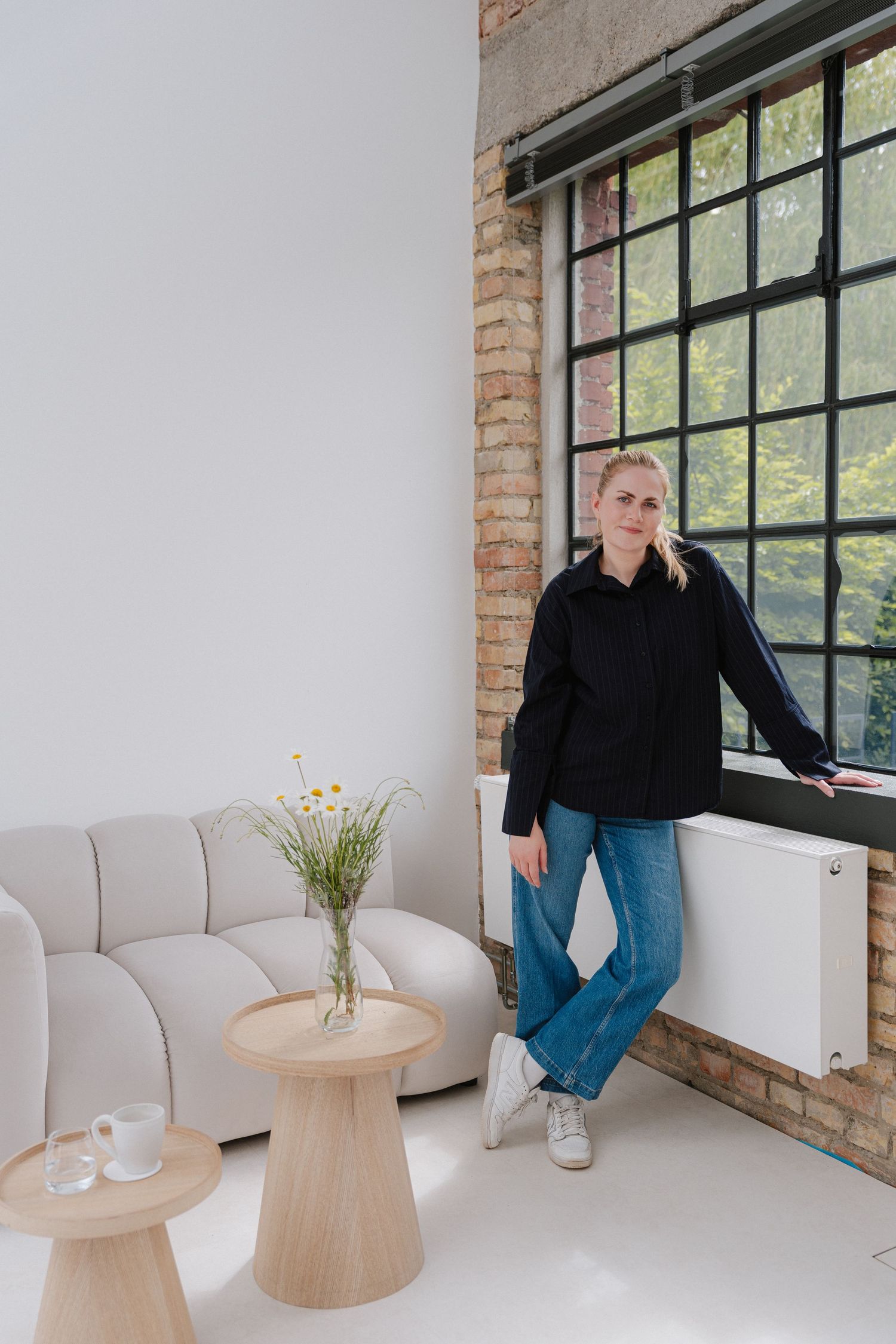 Woman in black shirt and jeans leaning against window in modern minimalist space with brick wall and neutral furniture.