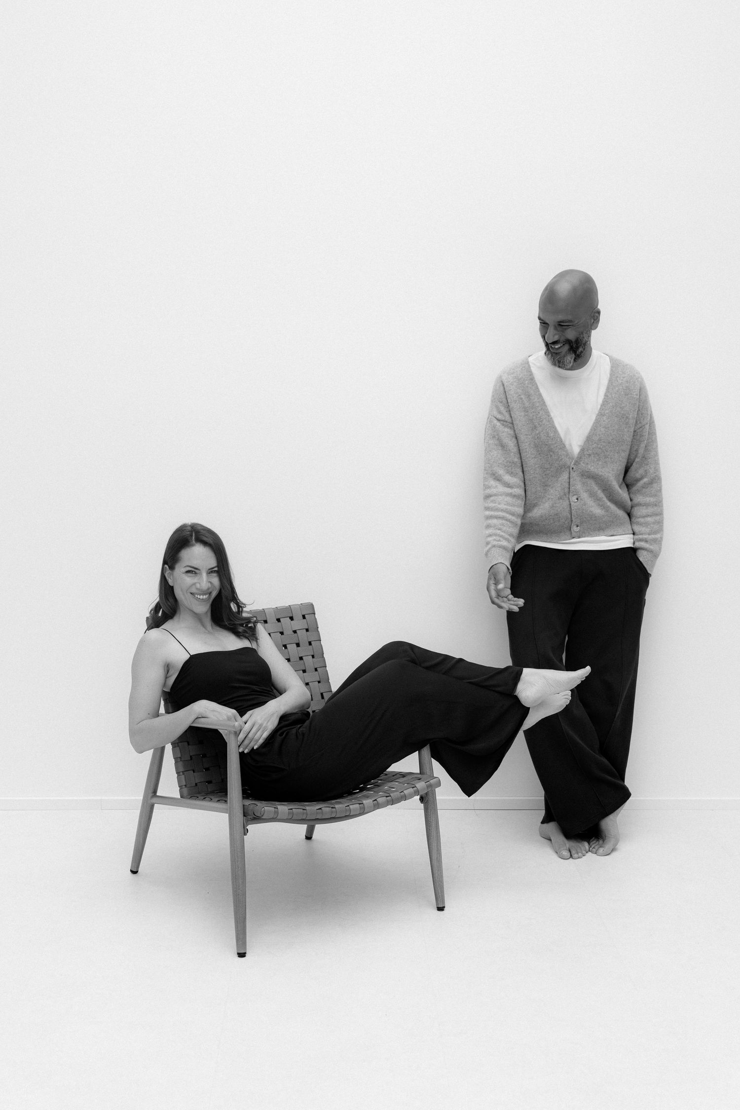 Black and white photo of a pregnant woman in a chair and a man standing beside her against a white wall.