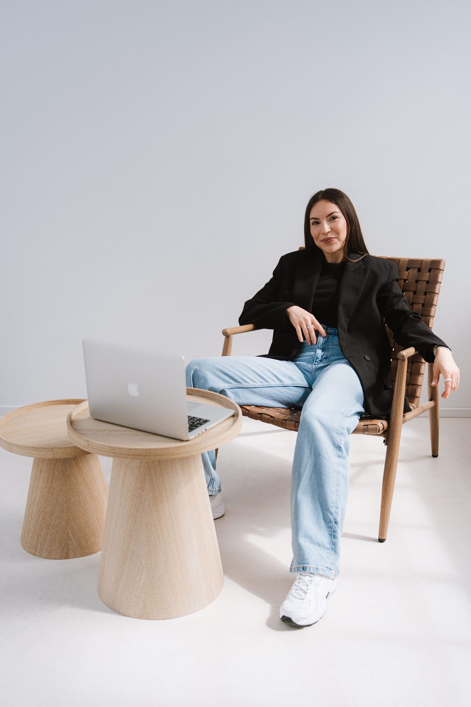 Woman in black blazer and jeans sits in wooden chair beside minimalist coffee table with open laptop.