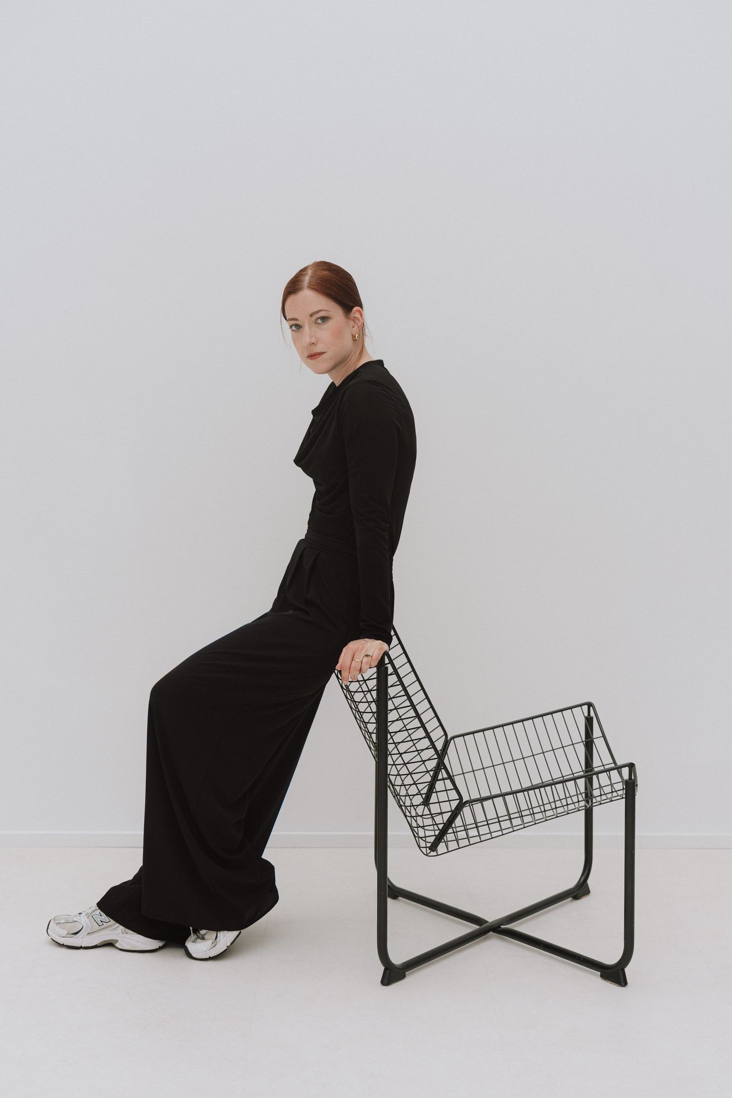 Woman in black wide-leg jumpsuit posing beside a minimalist wire chair against a white background.