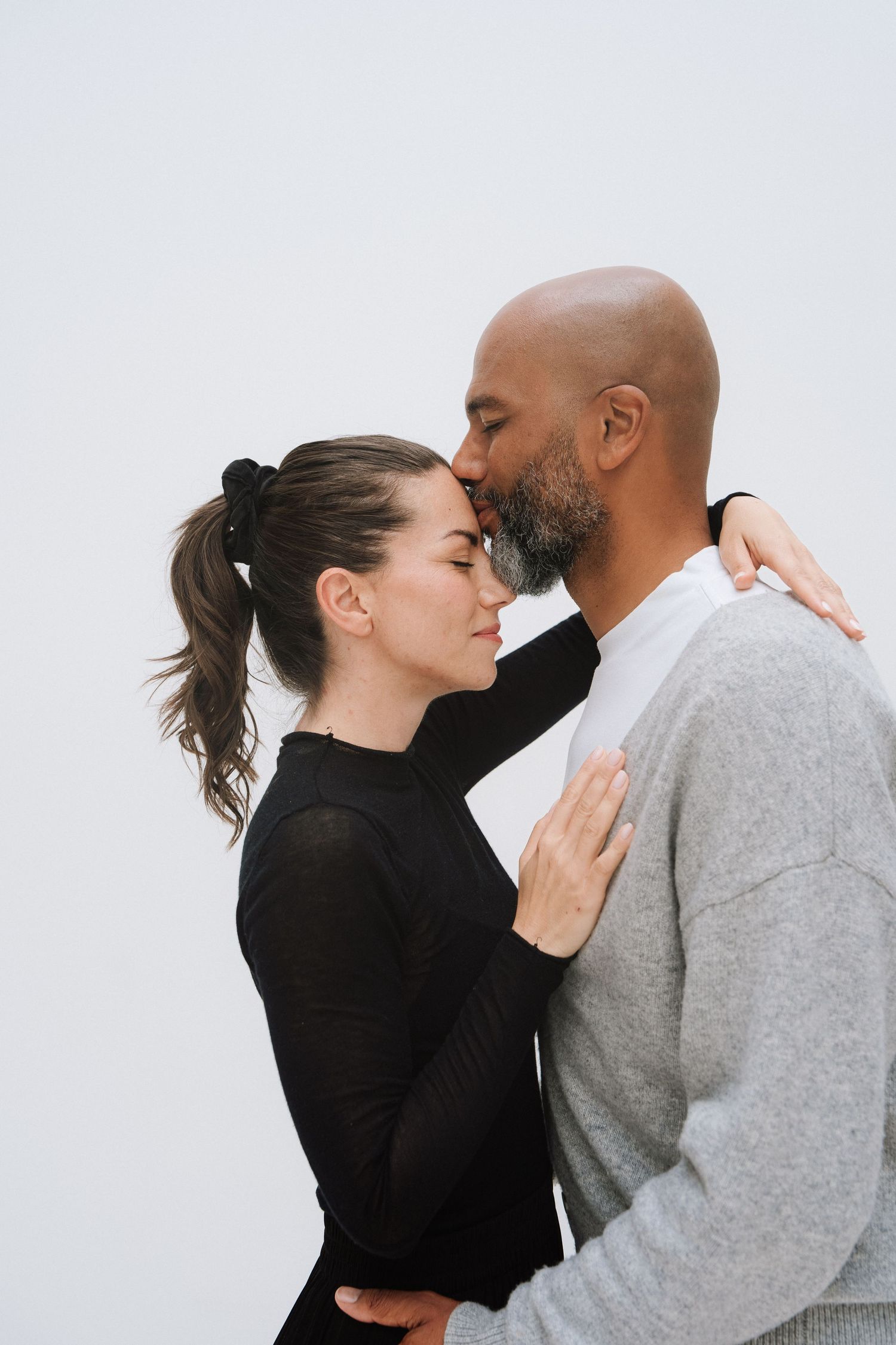A tender interracial couple sharing a kiss, woman in black turtleneck embracing bald man in gray sweater against white background.