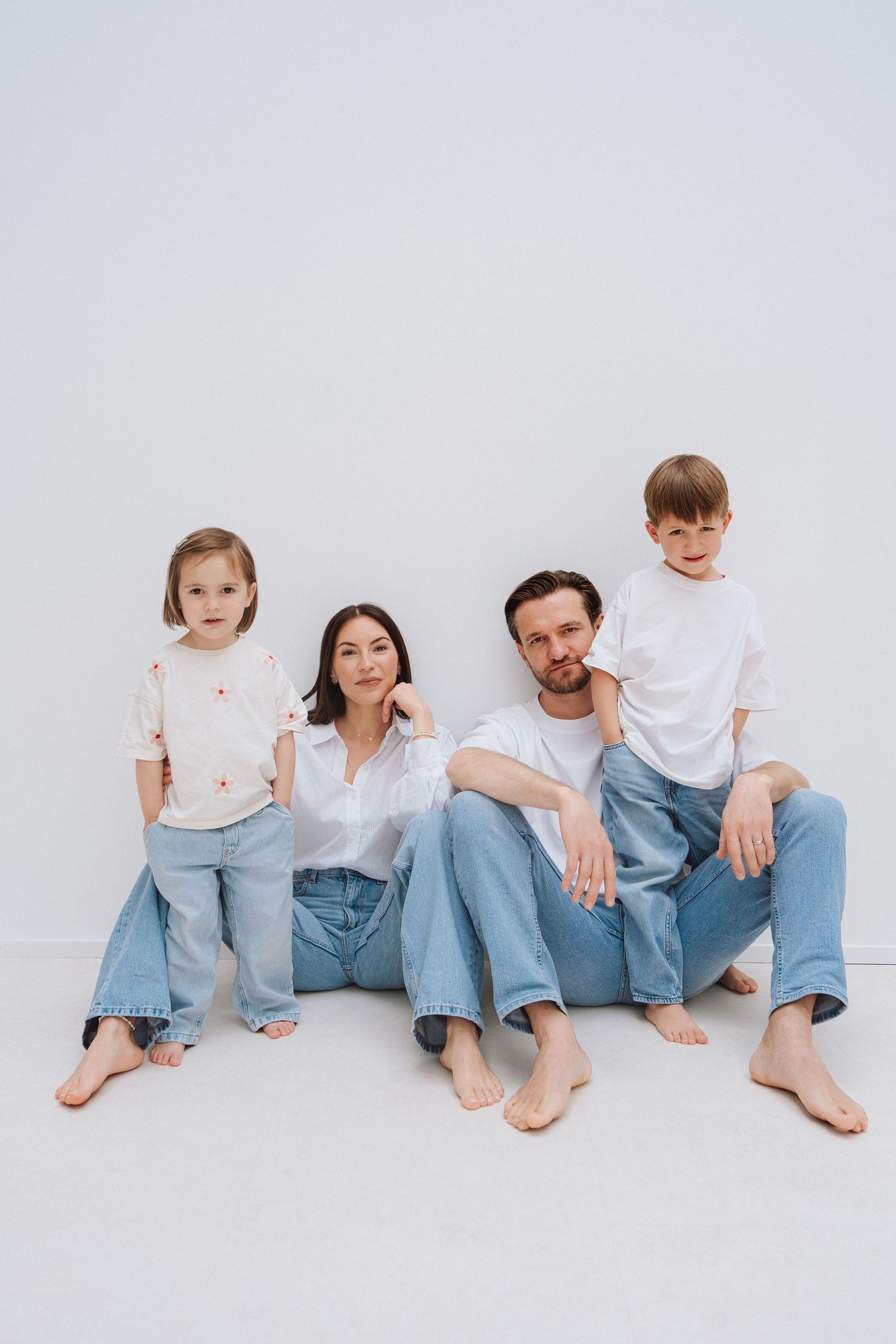Family of four in matching white shirts and jeans sitting together barefoot on a white studio floor.