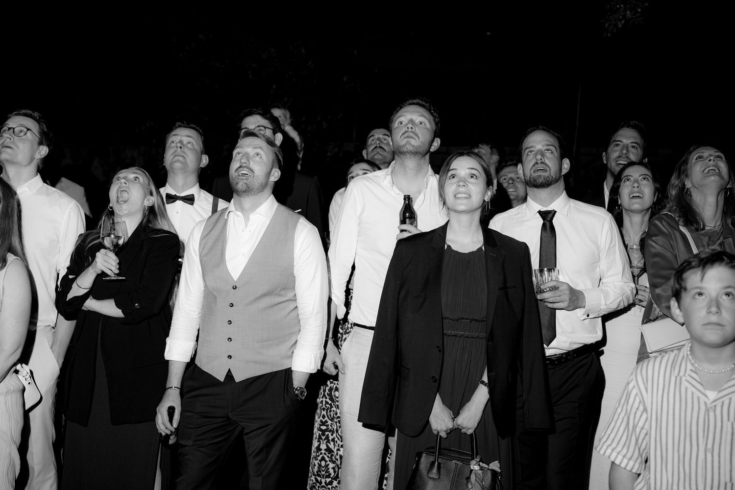 Large group of wedding guests dancing and celebrating at reception in dark venue.