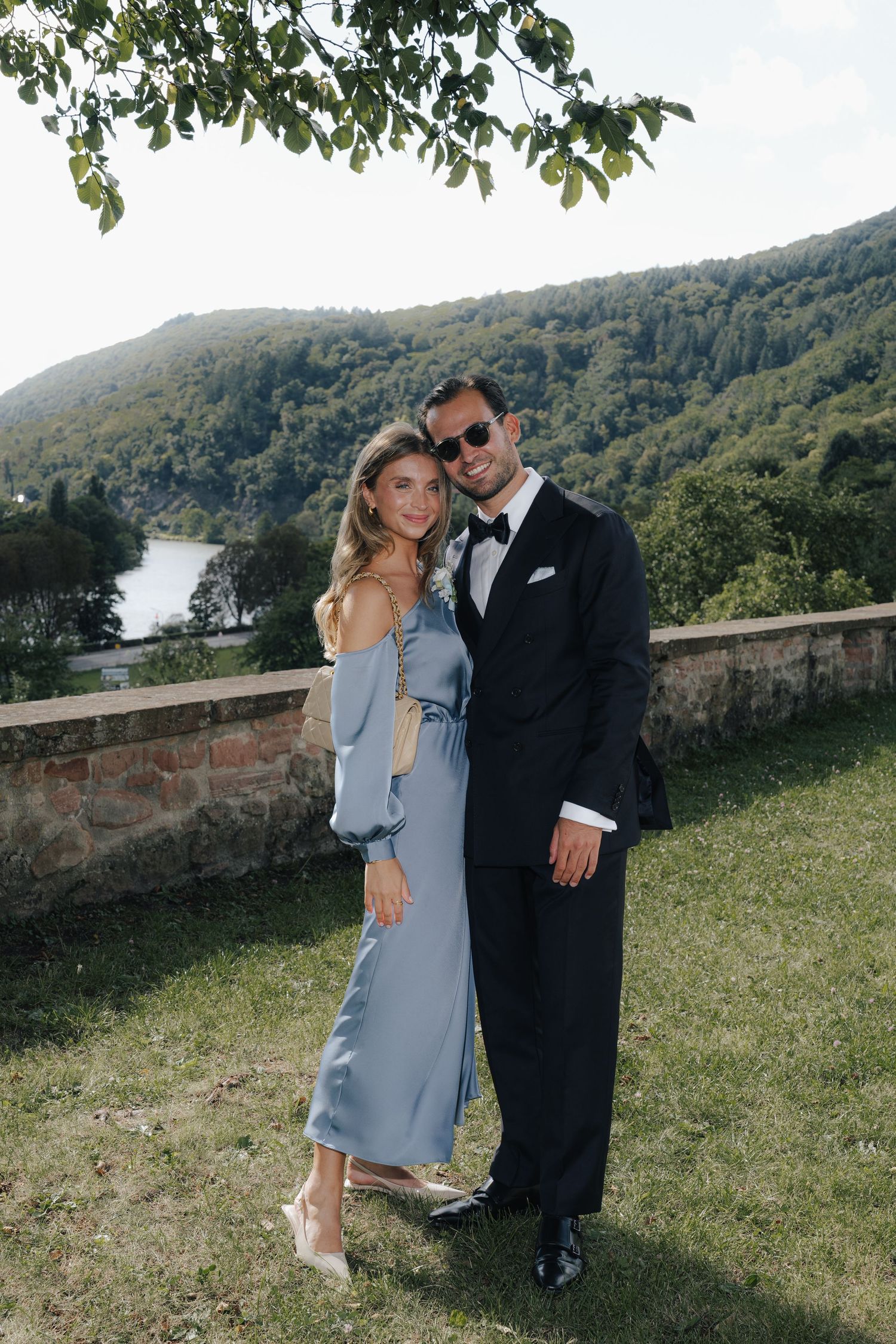 Couple in formal attire posing on a scenic overlook with mountains and water in the background.