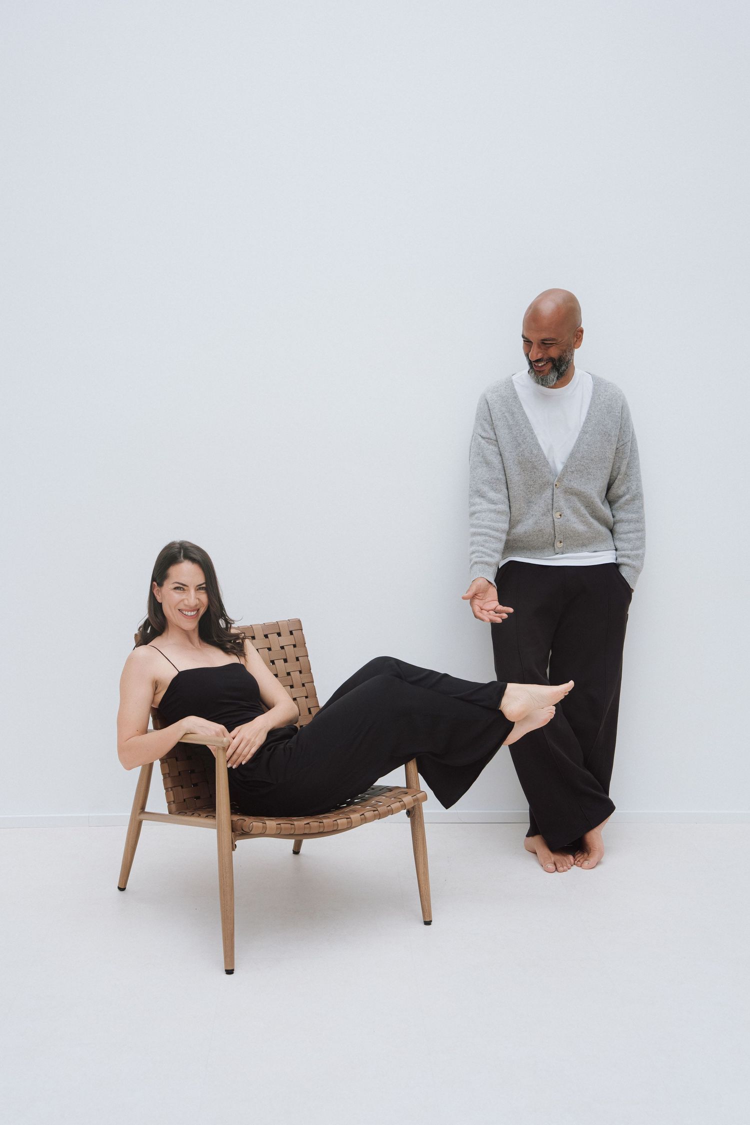 A woman in black lounges in a woven chair while a man in gray cardigan stands beside her against a white wall.