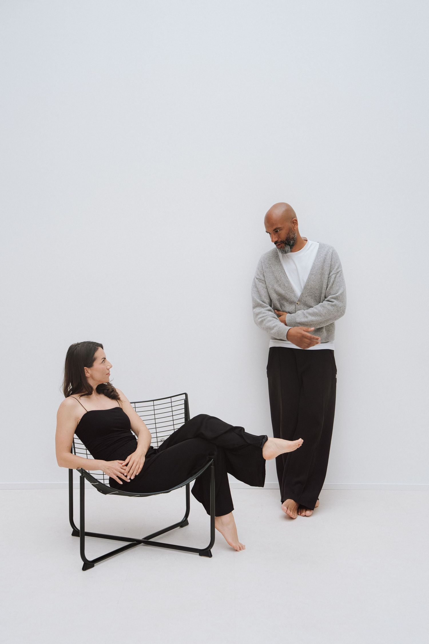 A woman relaxes in a black wire chair while a man in a gray cardigan stands nearby against a white background.