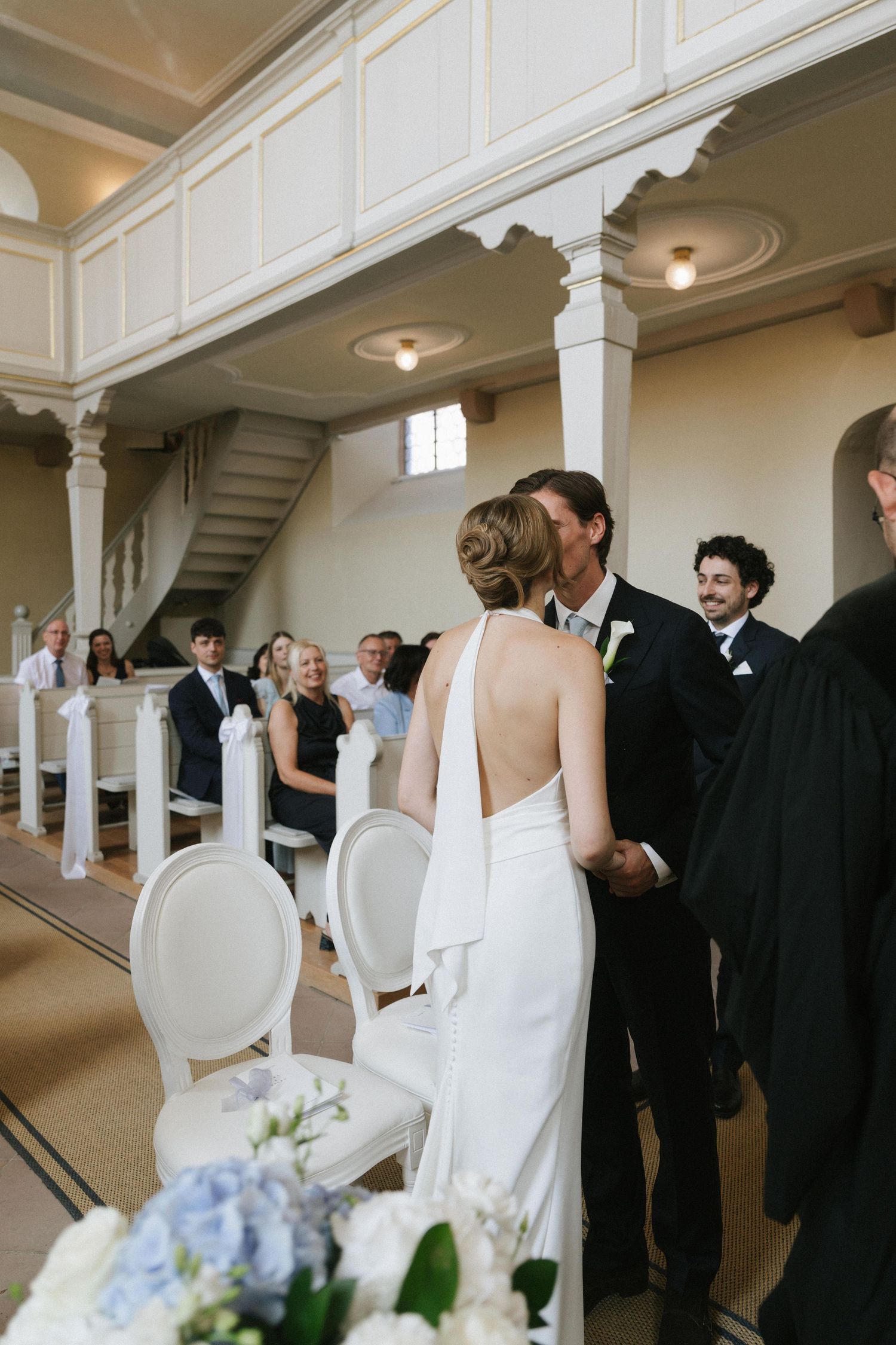Bride in backless white gown holds groom's hands at church altar during wedding ceremony with guests seated in pews.