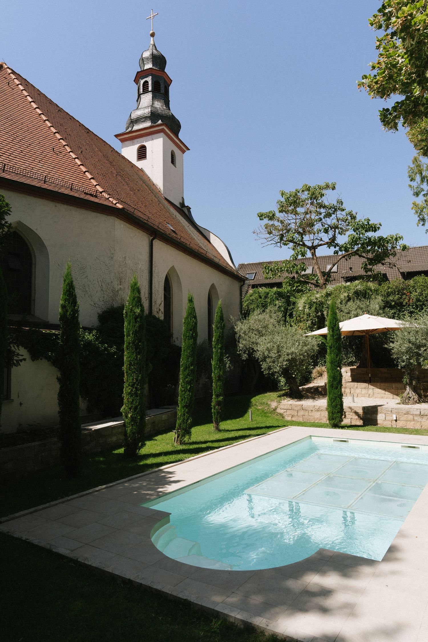 Historic European church with bell tower beside a serene outdoor swimming pool, surrounded by cypress trees and lush gardens.
