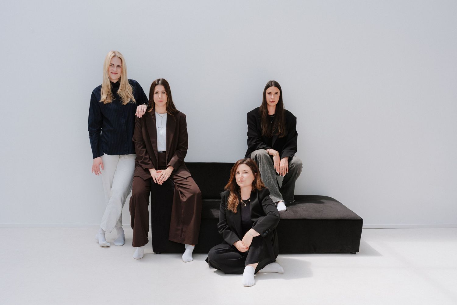 Four women in stylish neutral outfits pose together in a minimalist studio setting with clean gray walls.