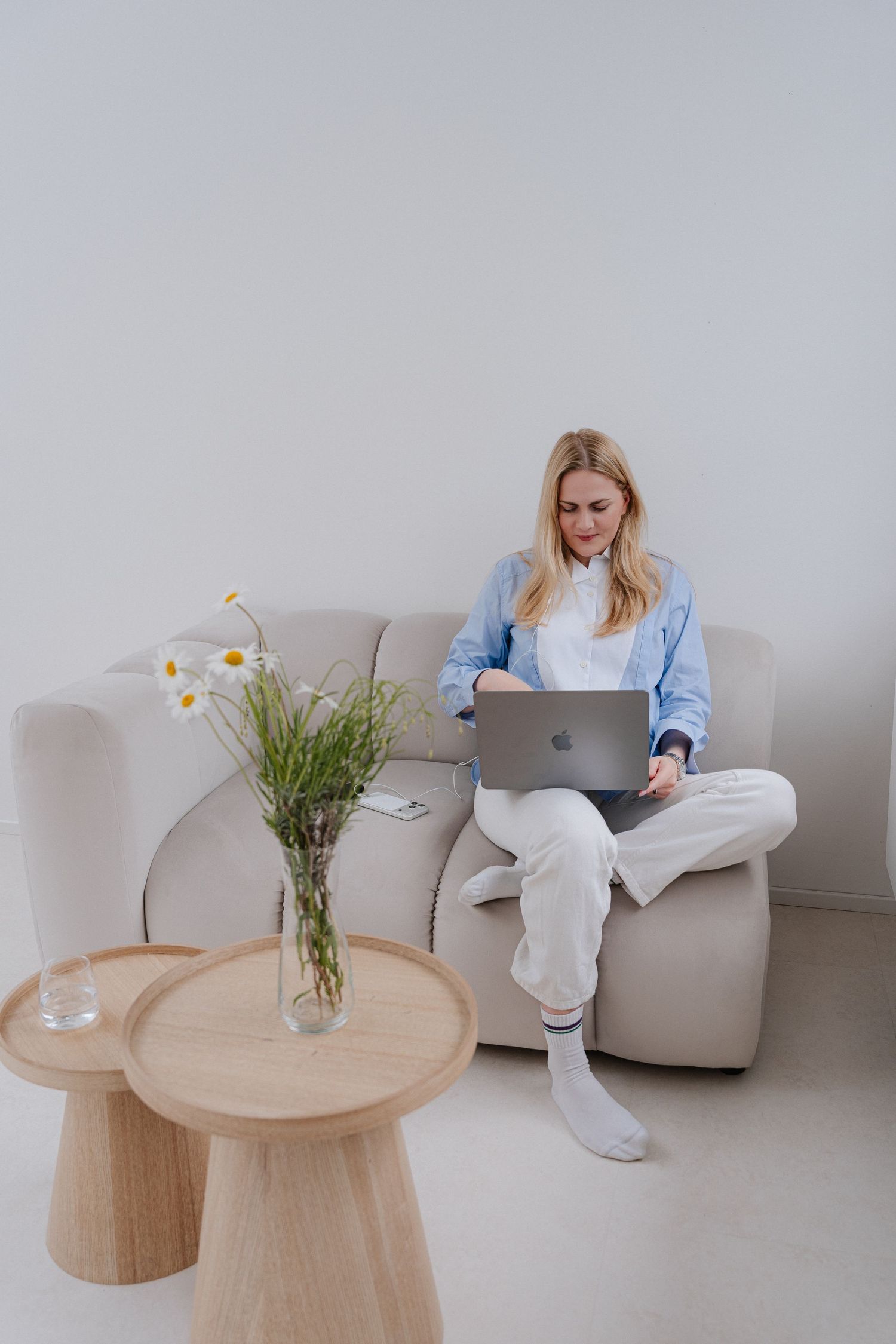Woman in casual blue shirt works on MacBook laptop while sitting on a beige sofa with daisy flowers on wooden table.