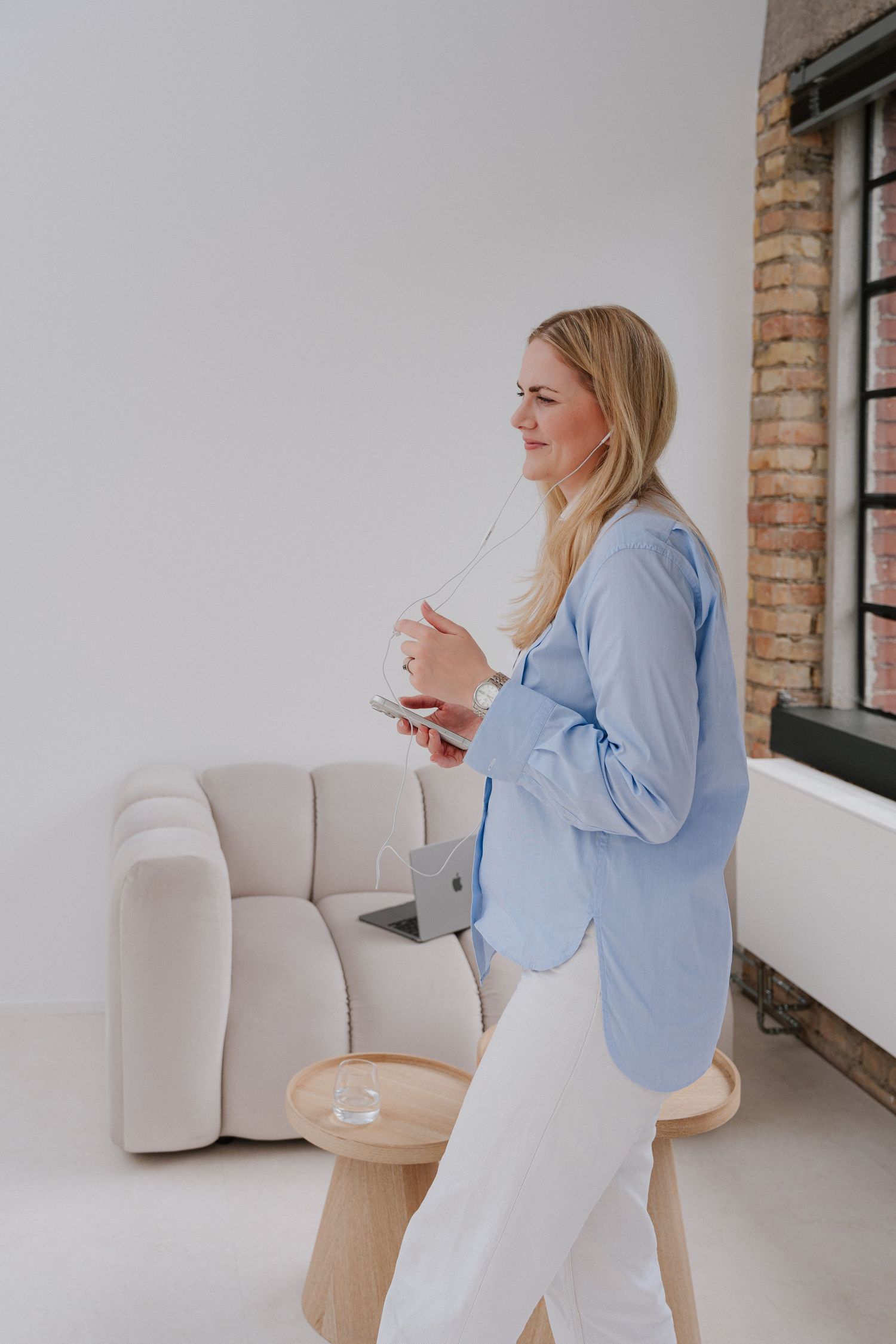 Woman in blue blazer and white pants holding a glass, standing in a modern minimalist white interior space.