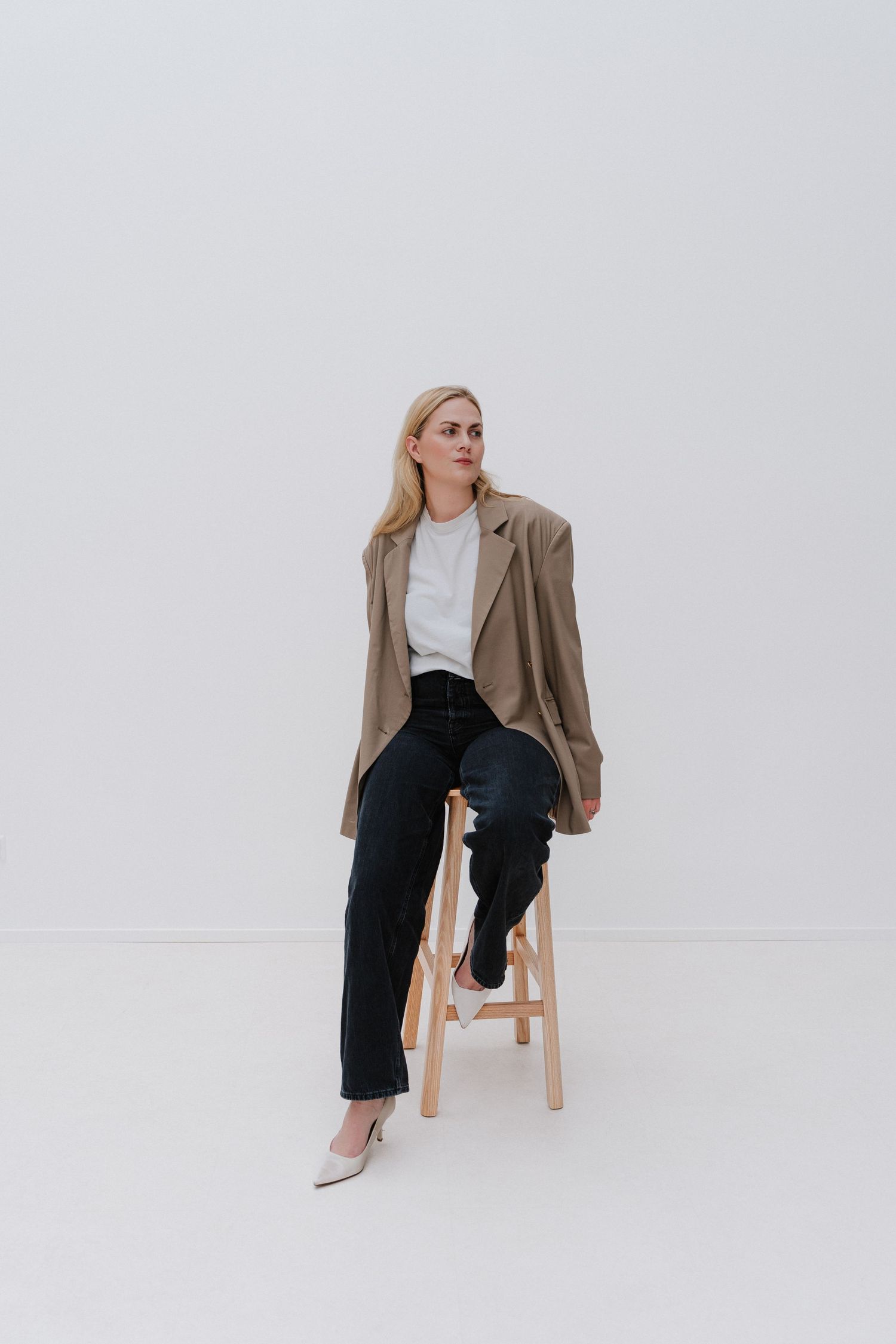 Blonde woman in beige blazer, white top, black jeans, and heels sitting on wooden stool against white background.