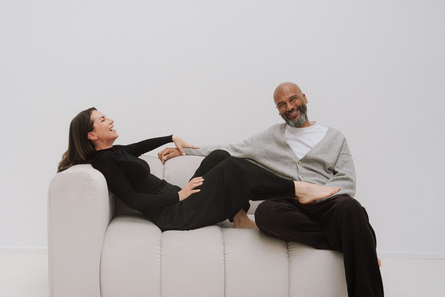 A man and woman relax on a white sofa, engaged in conversation against a minimalist white background.