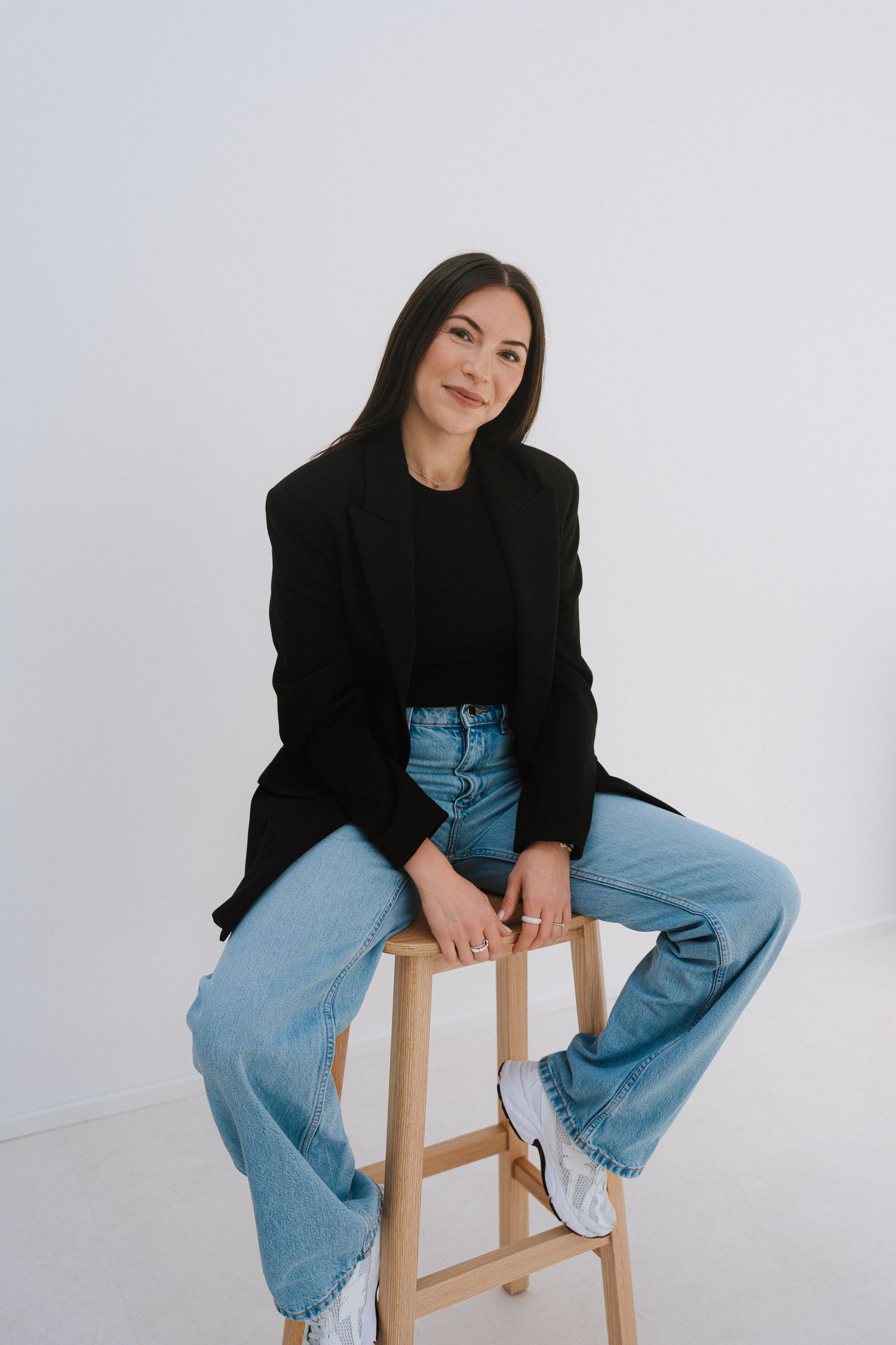 Woman in black blazer and jeans sitting on wooden stool against white background, smiling confidently.