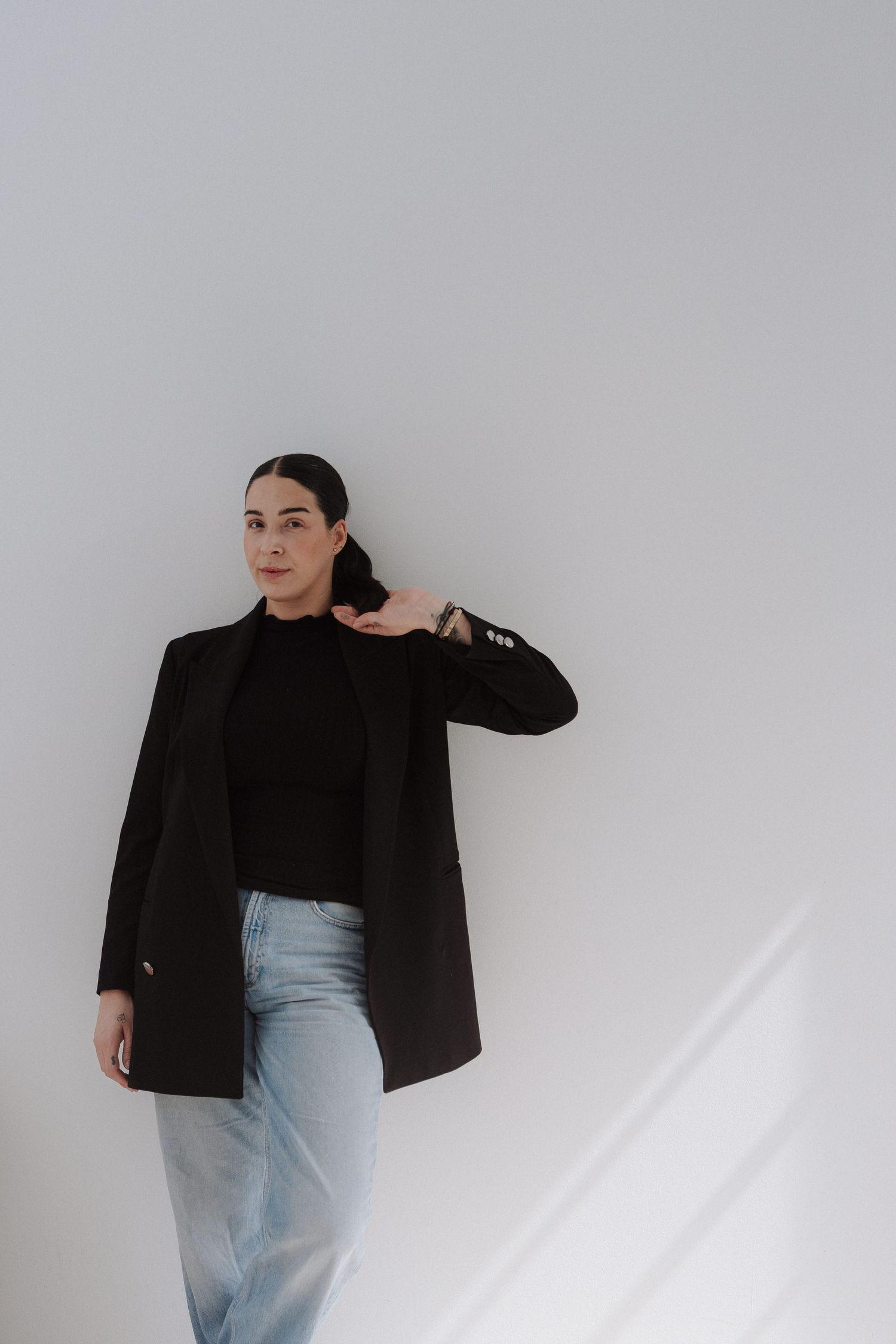 Woman in black blazer and jeans posing confidently against a white wall, wearing a black turtleneck and watch.