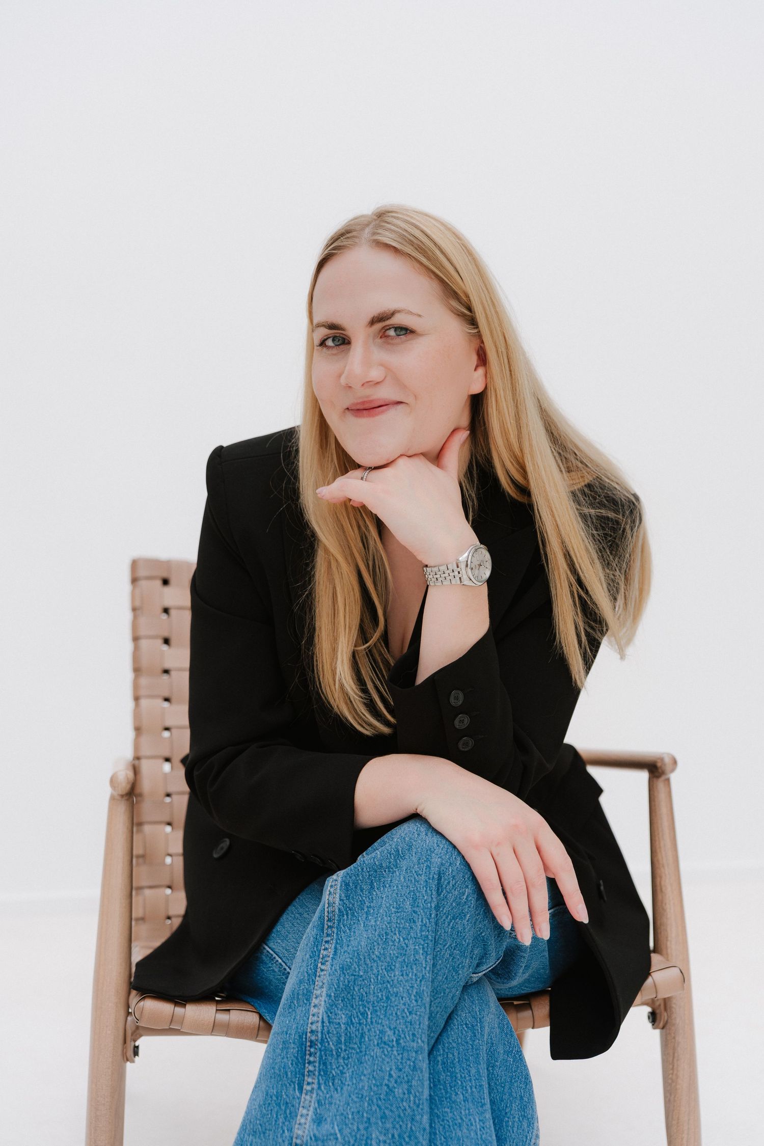 Blonde woman in black blazer and jeans sits relaxed on wooden chair, wearing silver watch, against white background.