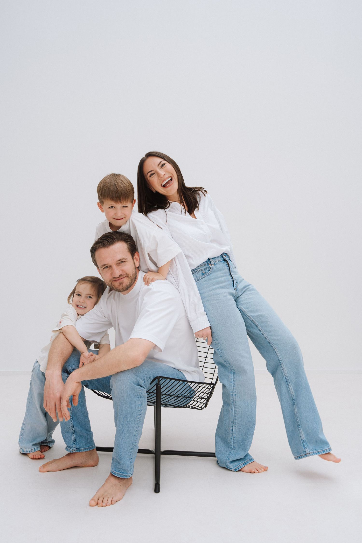 Happy family of four in white tees and jeans posing together on a chair against a white background.
