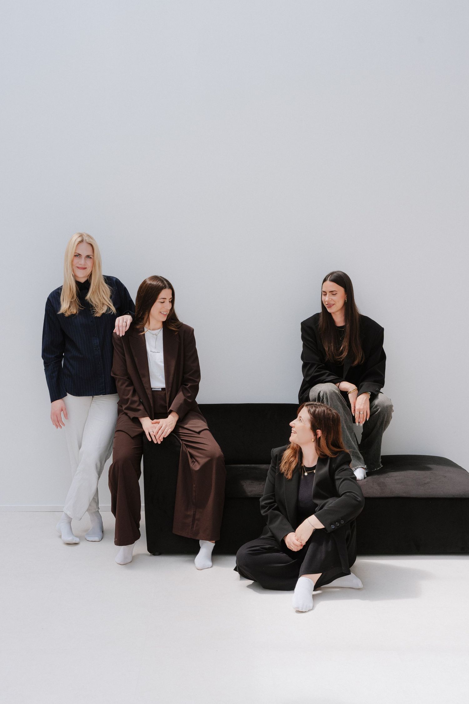 Four women in stylish dark outfits pose together near a black bench against a light gray background.