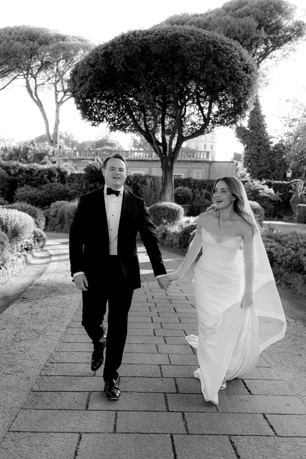 Bride and groom walk hand in hand through elegant garden path in black and white wedding portrait.