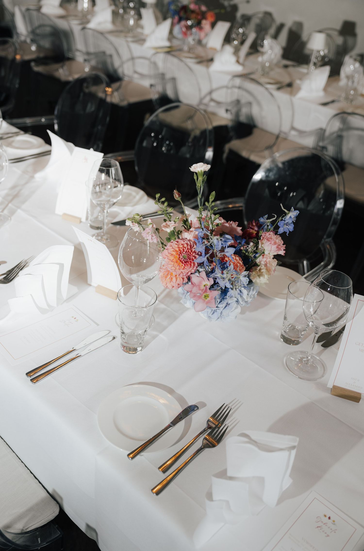 Wedding reception table setting with white linens, gold cutlery and delicate pink and blue floral centerpieces.