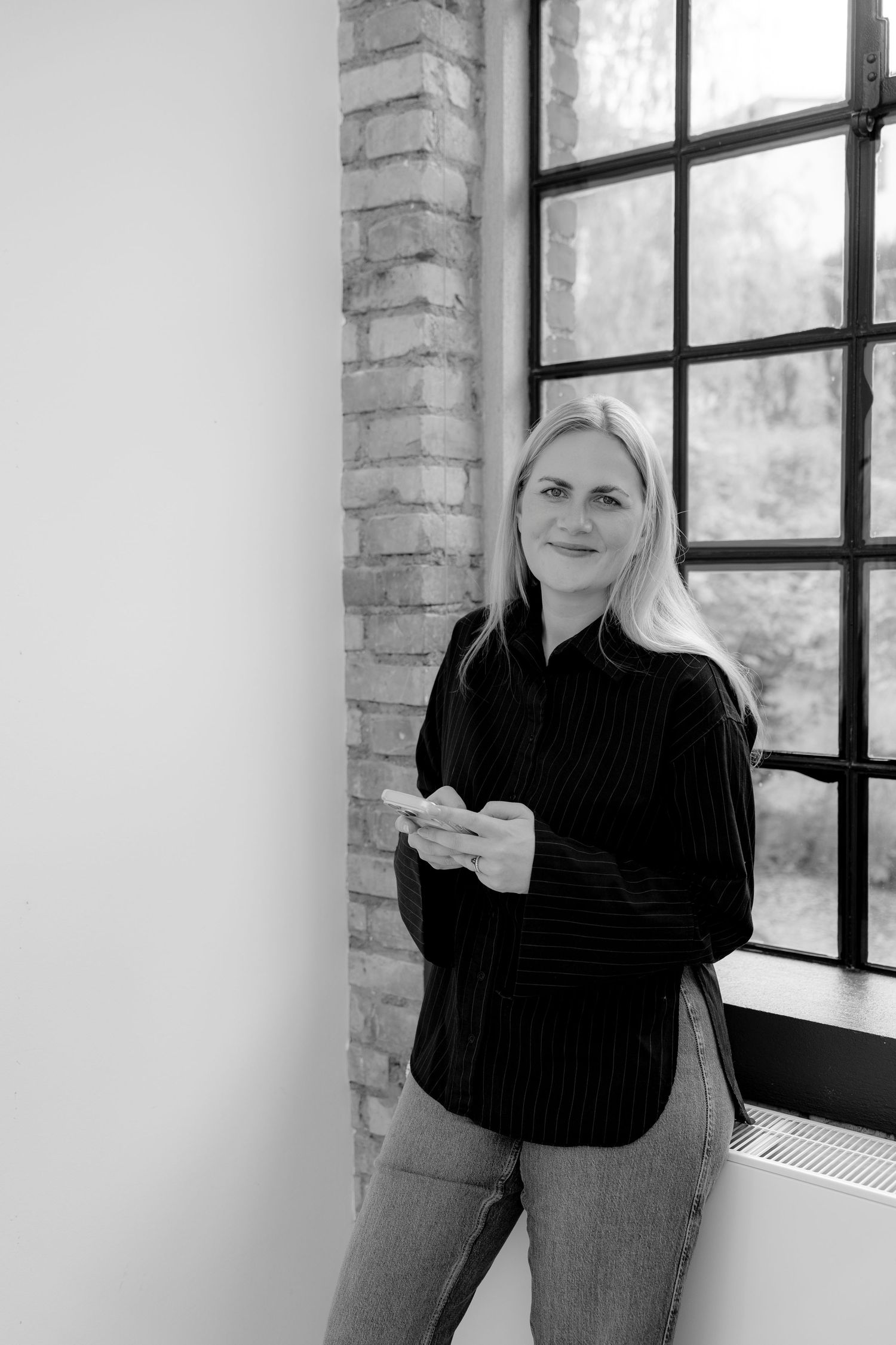 Black and white photo of a blonde woman in a black shirt holding a phone, leaning against a brick wall by a large window.