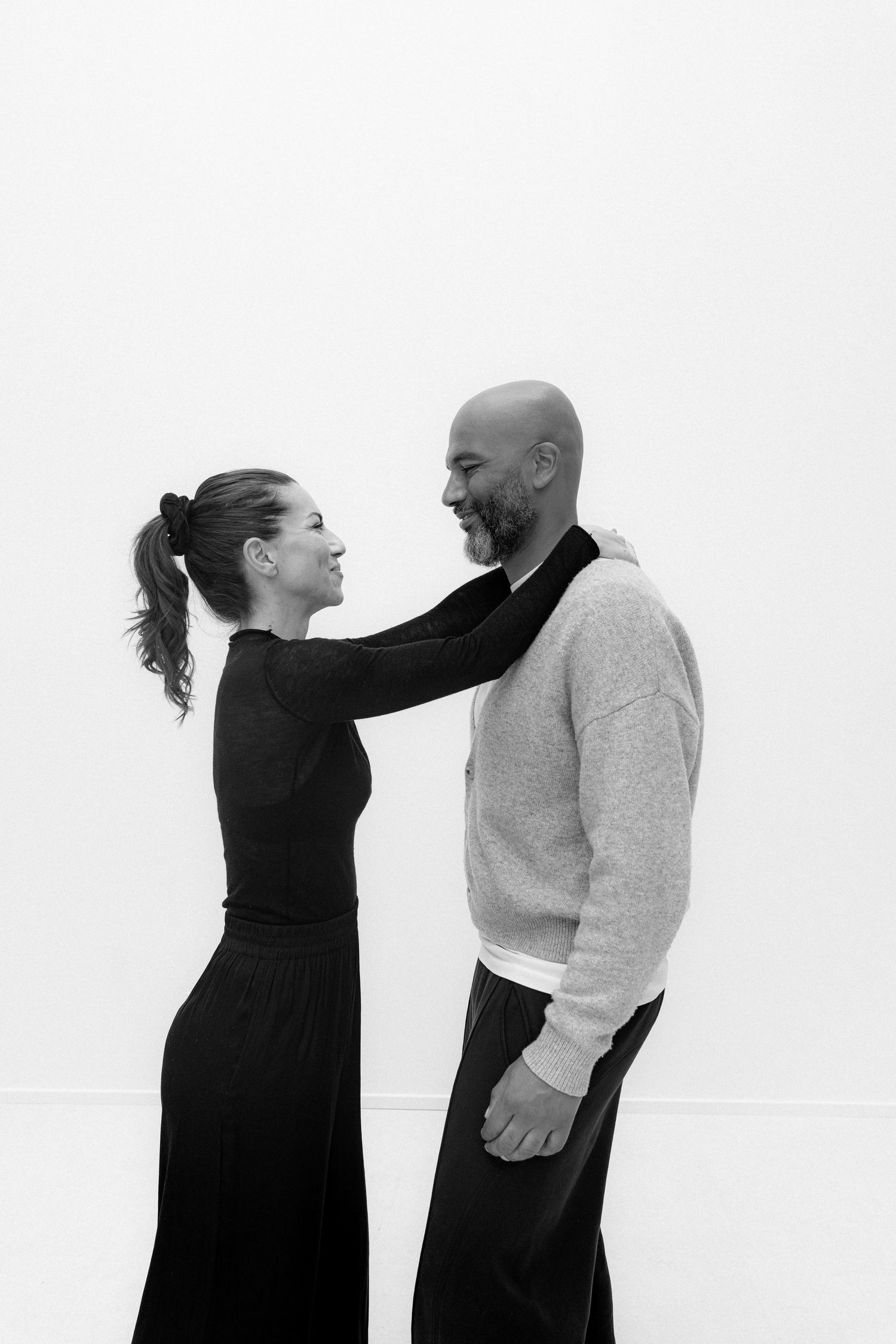 Black and white photo of a smiling couple facing each other, woman in black outfit resting hand on mans shoulder.