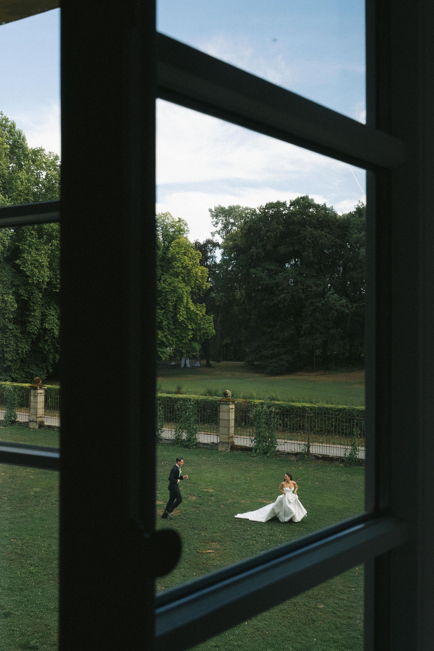 Bride and groom photographed through a window in a lush green garden during their wedding day outdoor photo session.