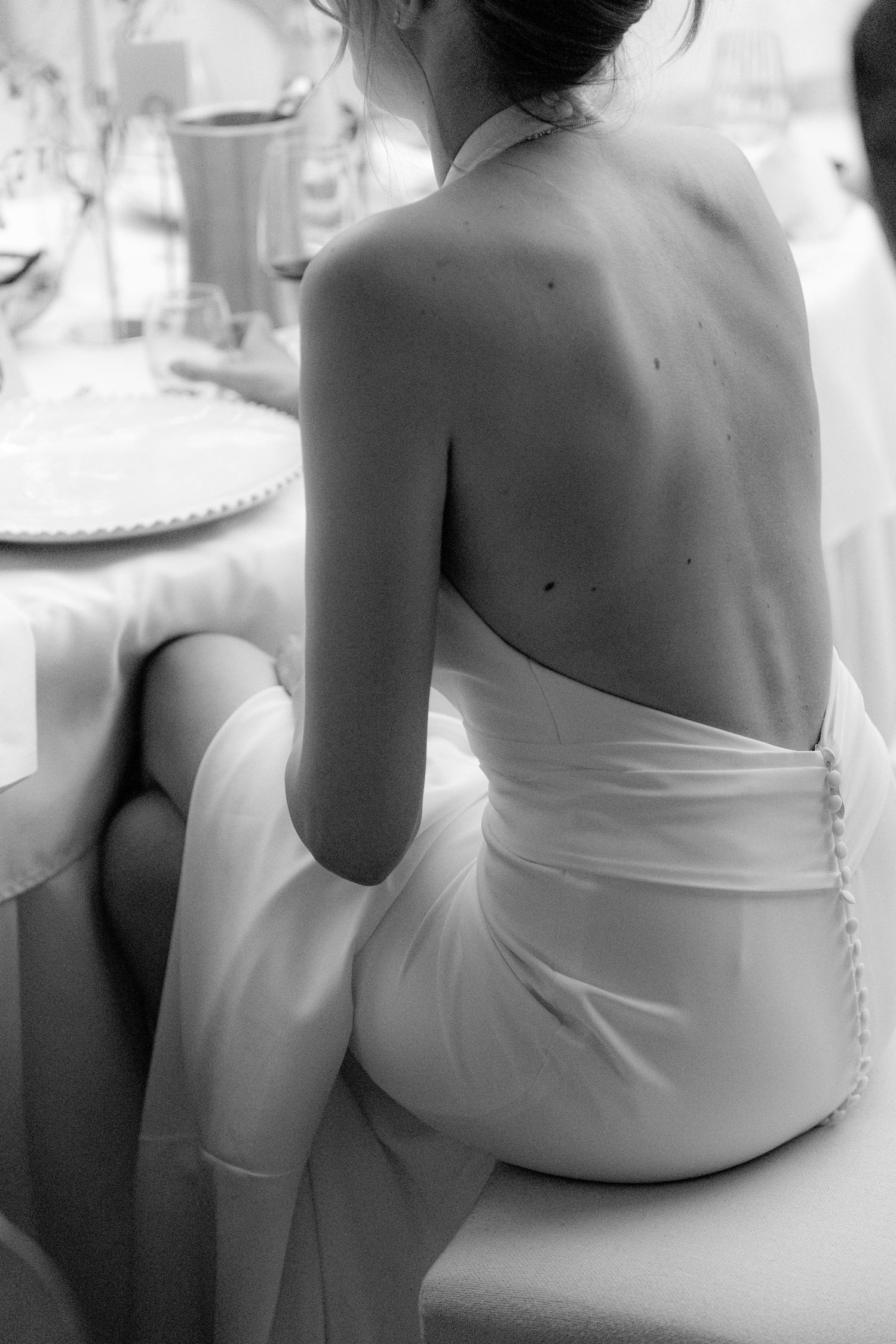 Black and white photo of a bride in a backless white halter wedding gown seated at an elegant reception table.