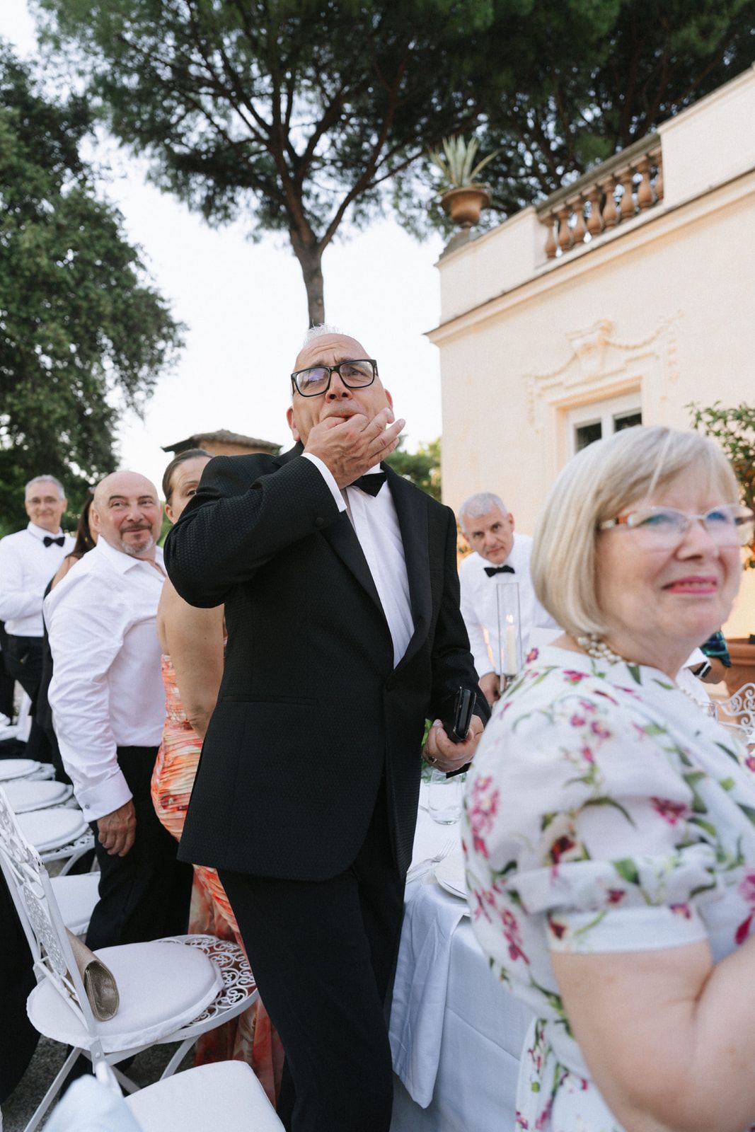 Elegantly dressed guests at an outdoor wedding reception near a classic villa, with a man in tuxedo gesturing expressively.