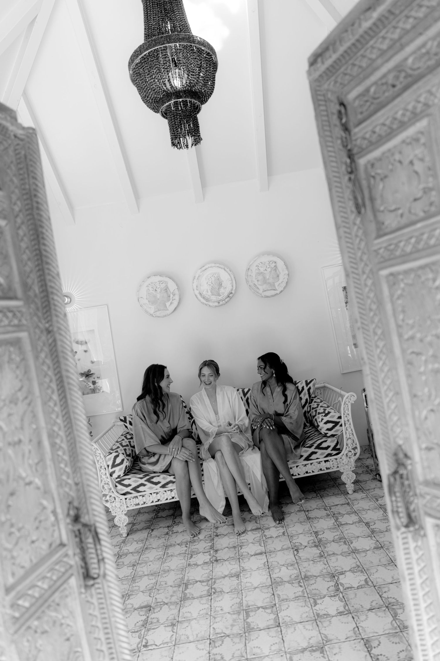 Three women in robes sit together on an ornate white sofa in an elegant Moroccan-style room, black and white photo.