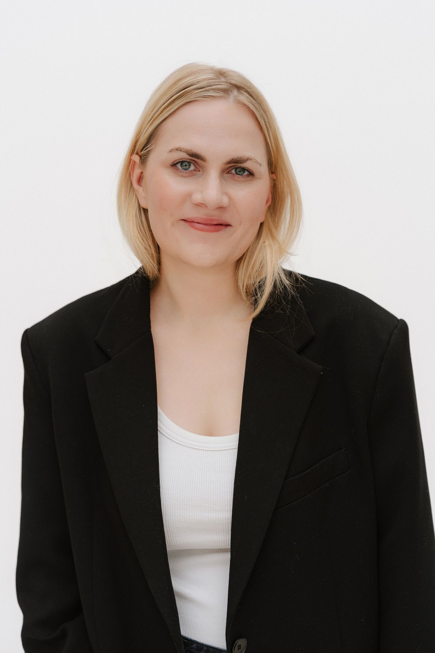 Professional blonde woman in black blazer and white top poses confidently against a white background.