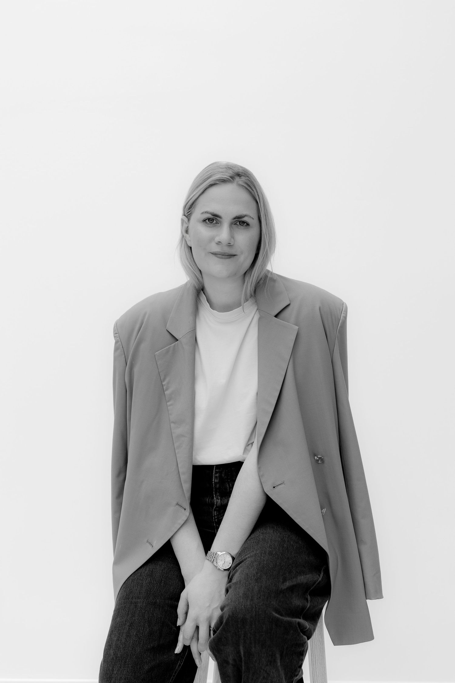 Black and white portrait of a blonde woman in a blazer and jeans, seated against a white background.