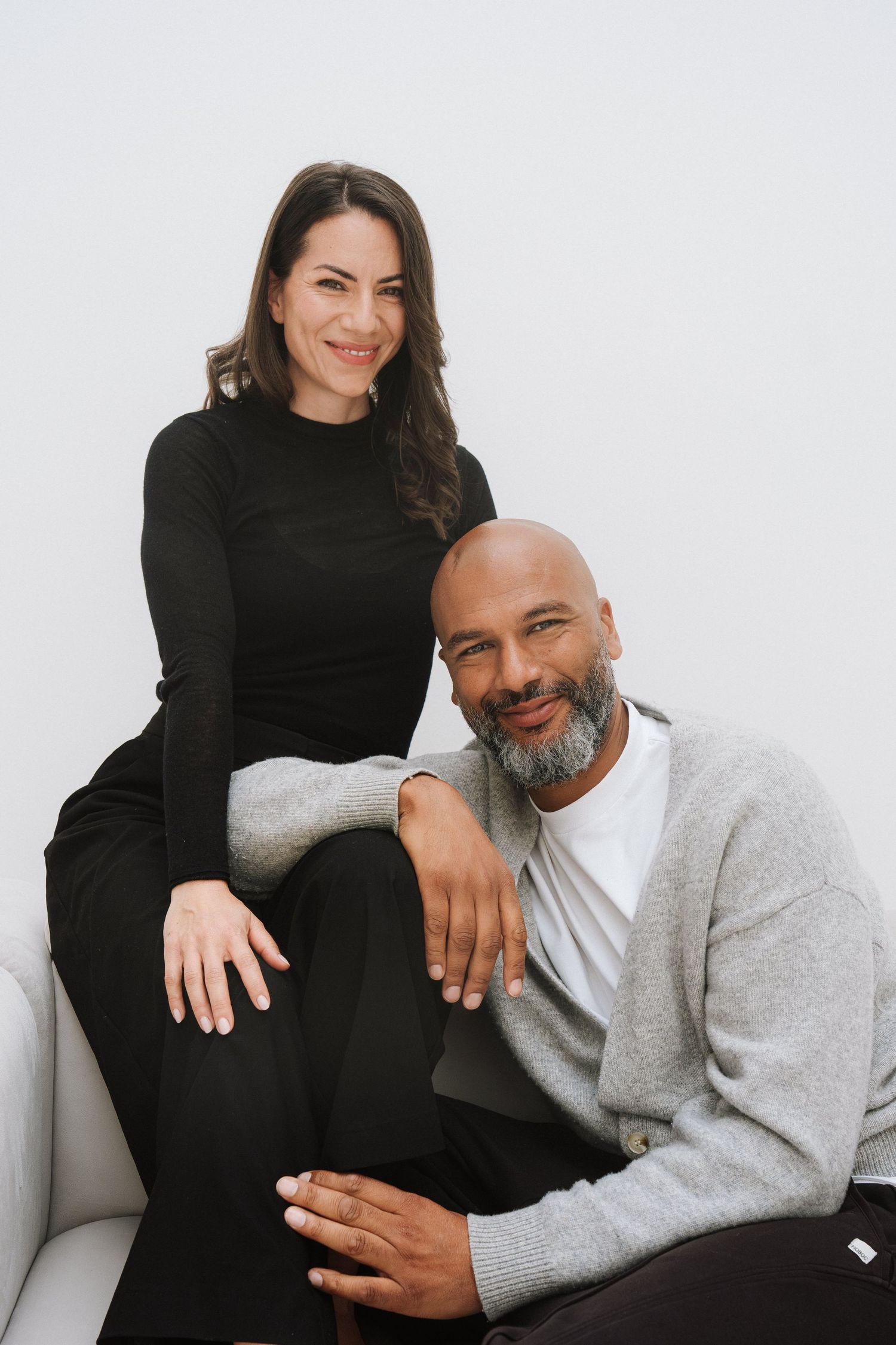 A smiling man and woman pose together on a white sofa, wearing casual black and gray outfits against a light background.