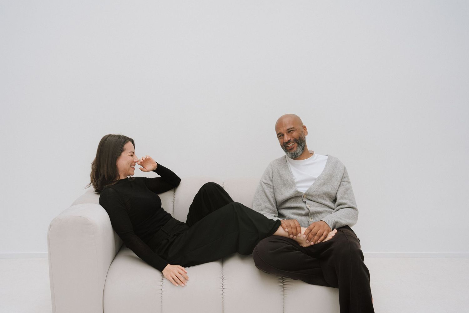 Two people relaxing on a white sofa, smiling and conversing in a bright, minimalist white room.
