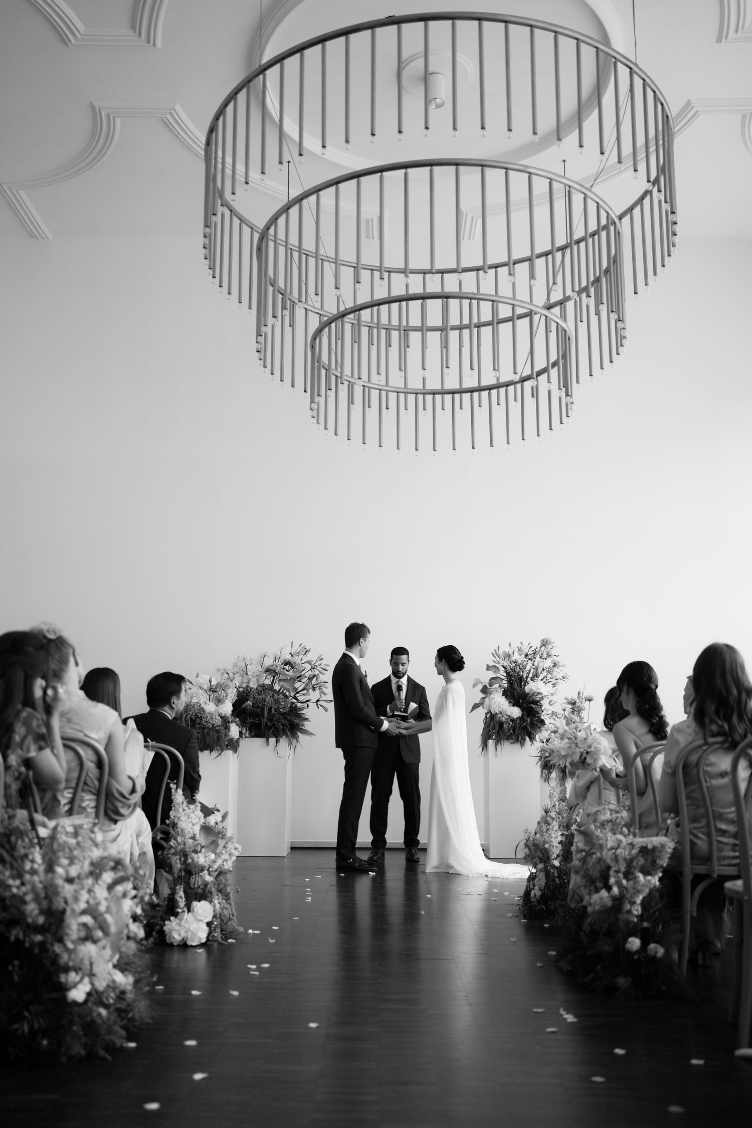 Wedding ceremony setup under modern circular chandeliers with floral arrangements lining the aisle.