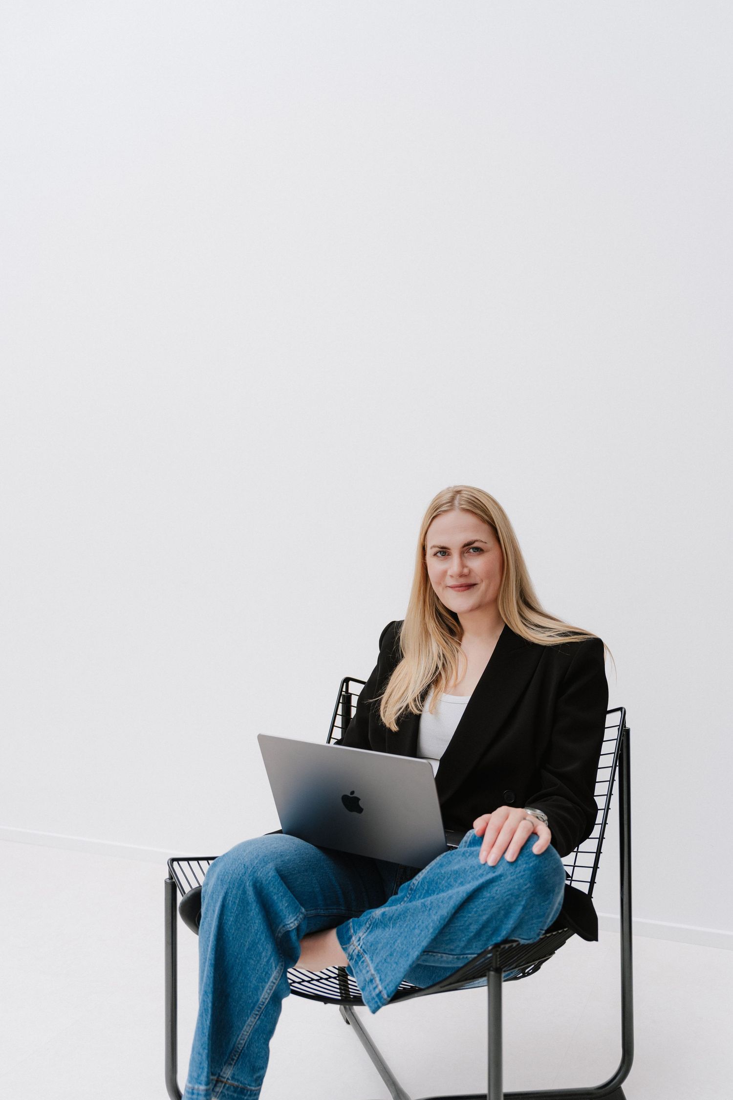 Woman in black blazer and jeans sitting on chair with Apple MacBook laptop against white background.