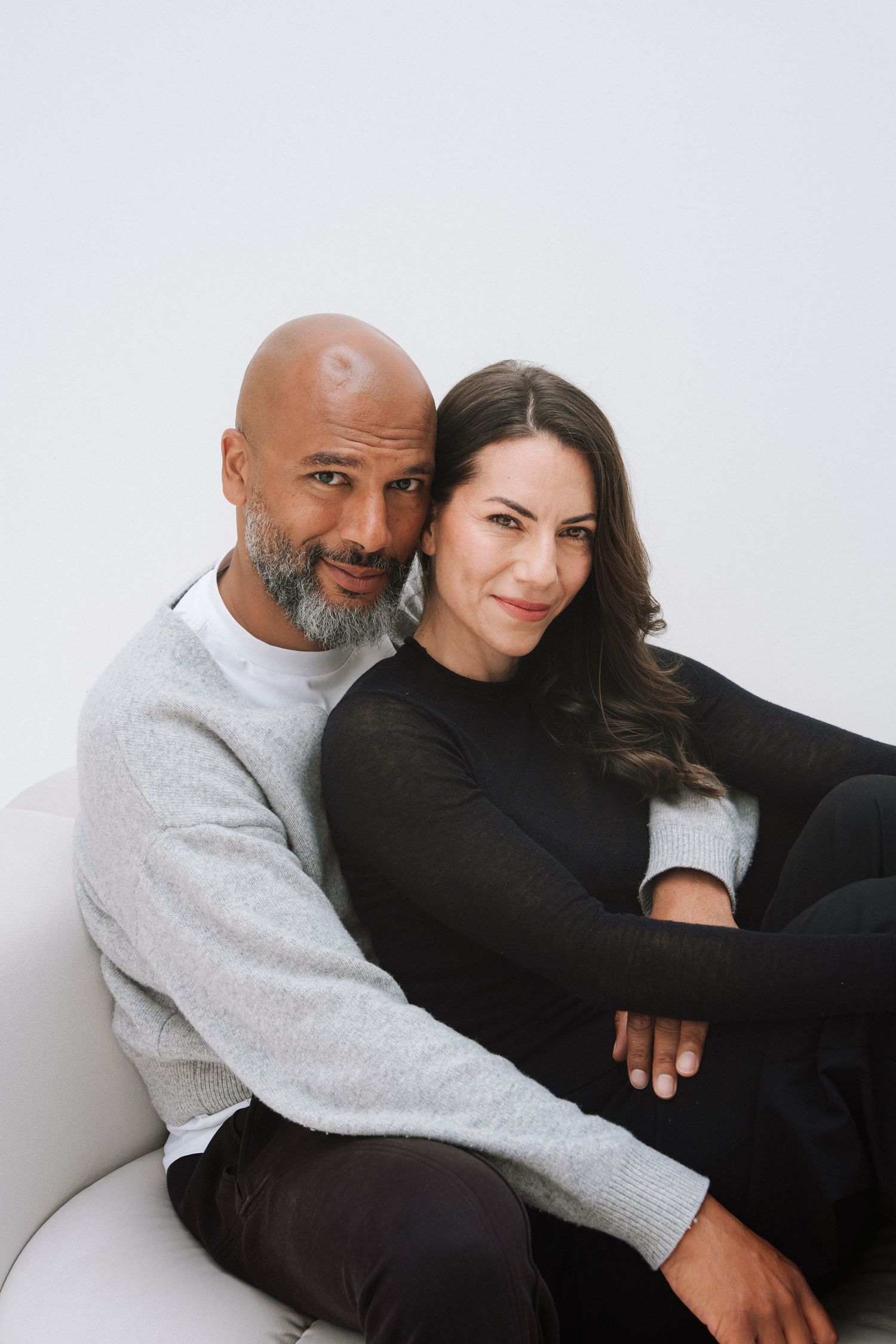 A bald man with a gray beard and a brunette woman sit closely together on a white couch, smiling warmly at the camera.