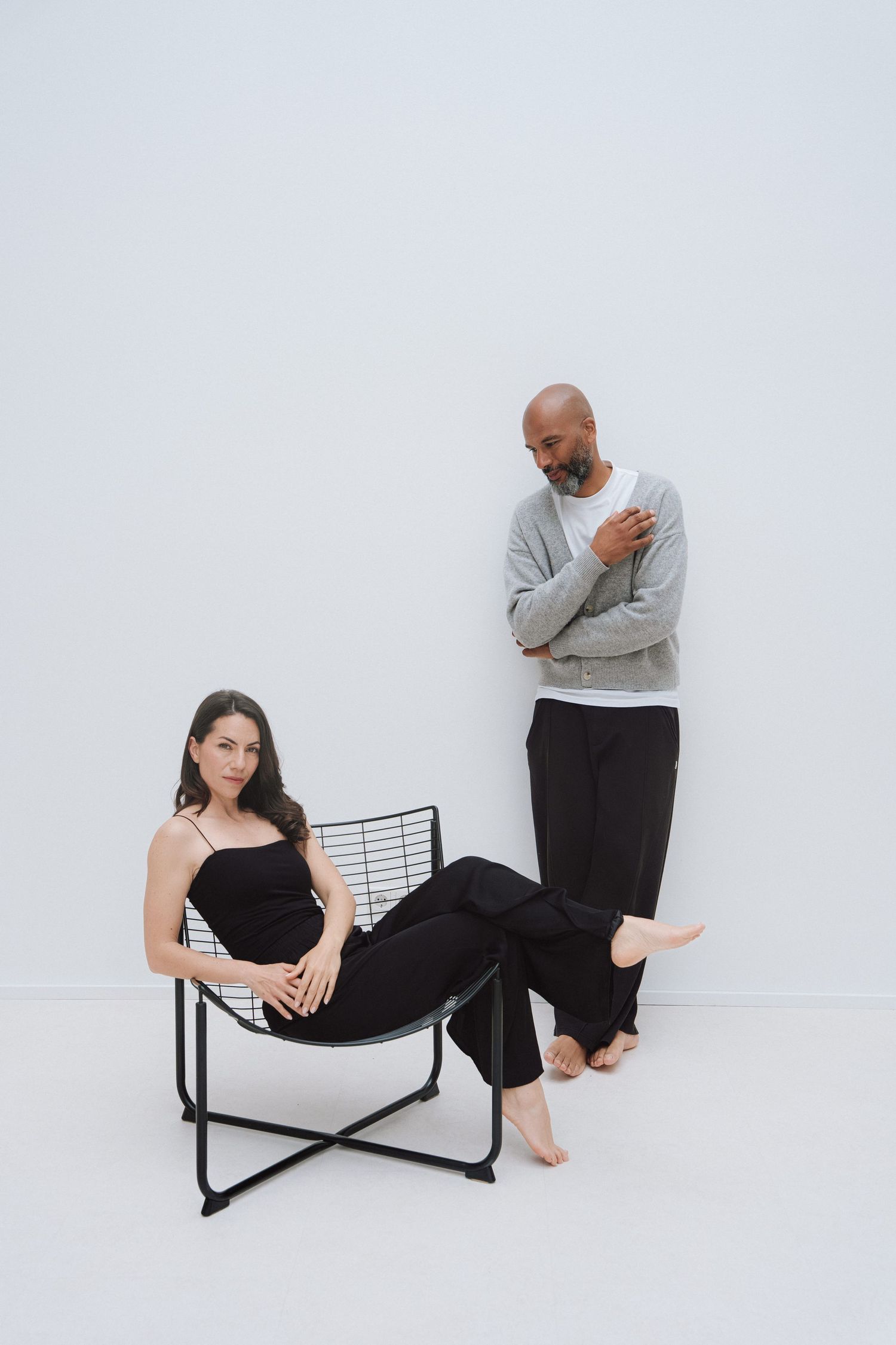 A woman in black reclines on a wire chair while a bald man in gray stands behind her against a white wall.