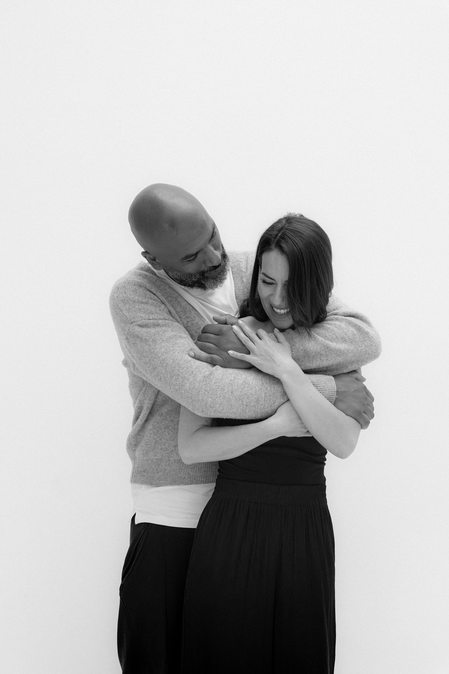 Black and white photo of a bald man with beard hugging a smiling woman in black skirt against white background.