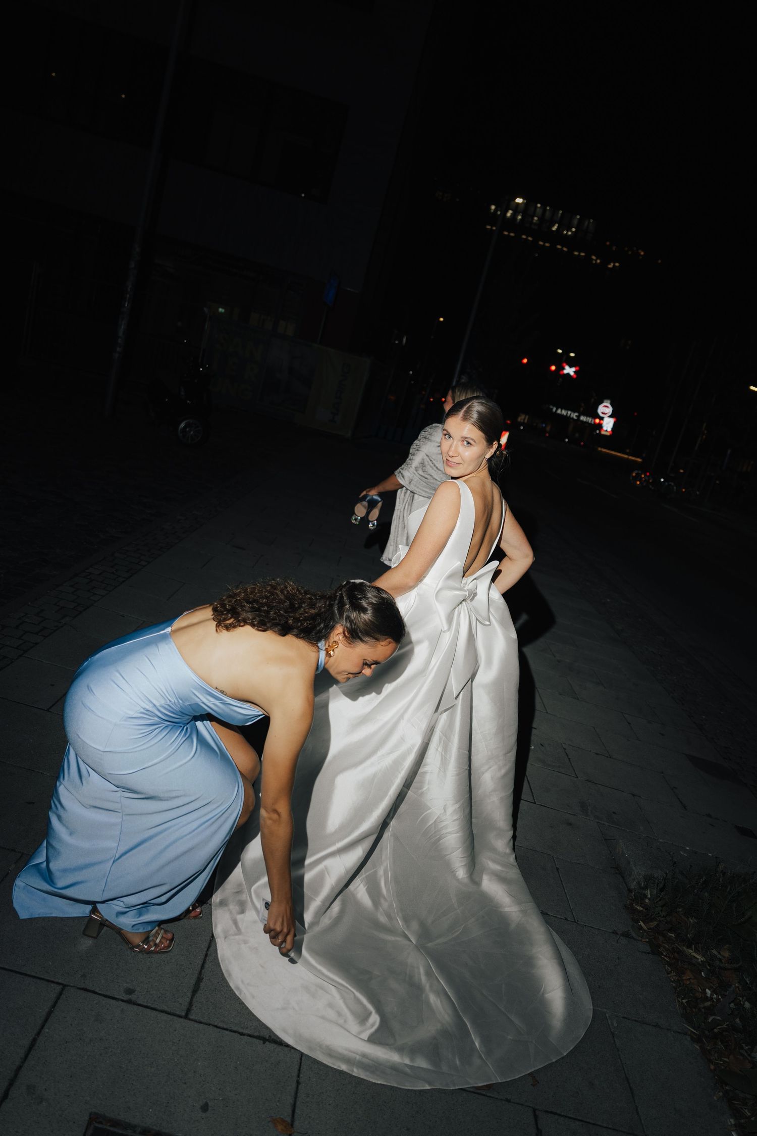 Person in blue dress helping adjust a white wedding dress train outdoors at night.