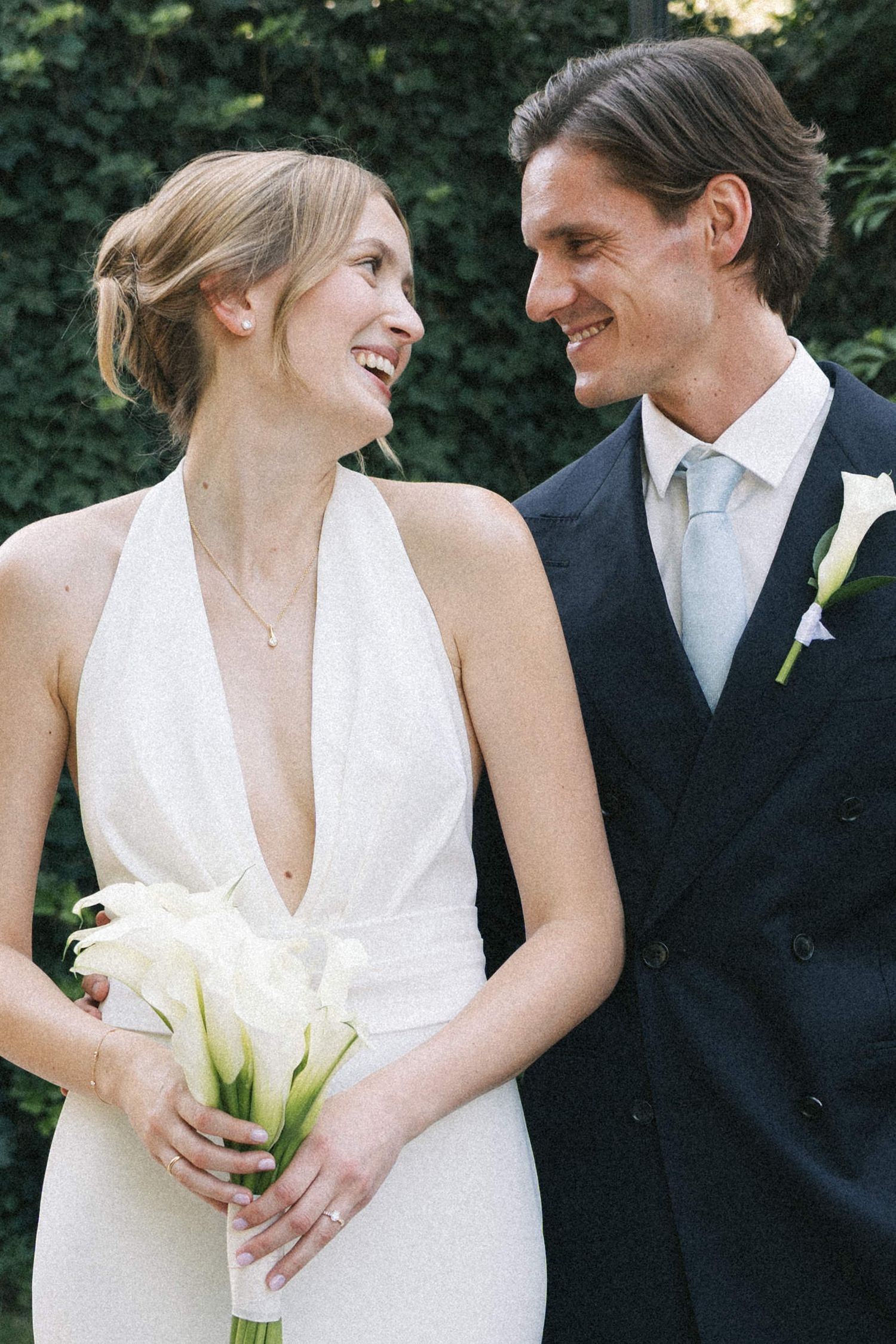 A smiling bride in a white halter gown holds calla lilies beside a groom in a navy suit with a light blue tie.