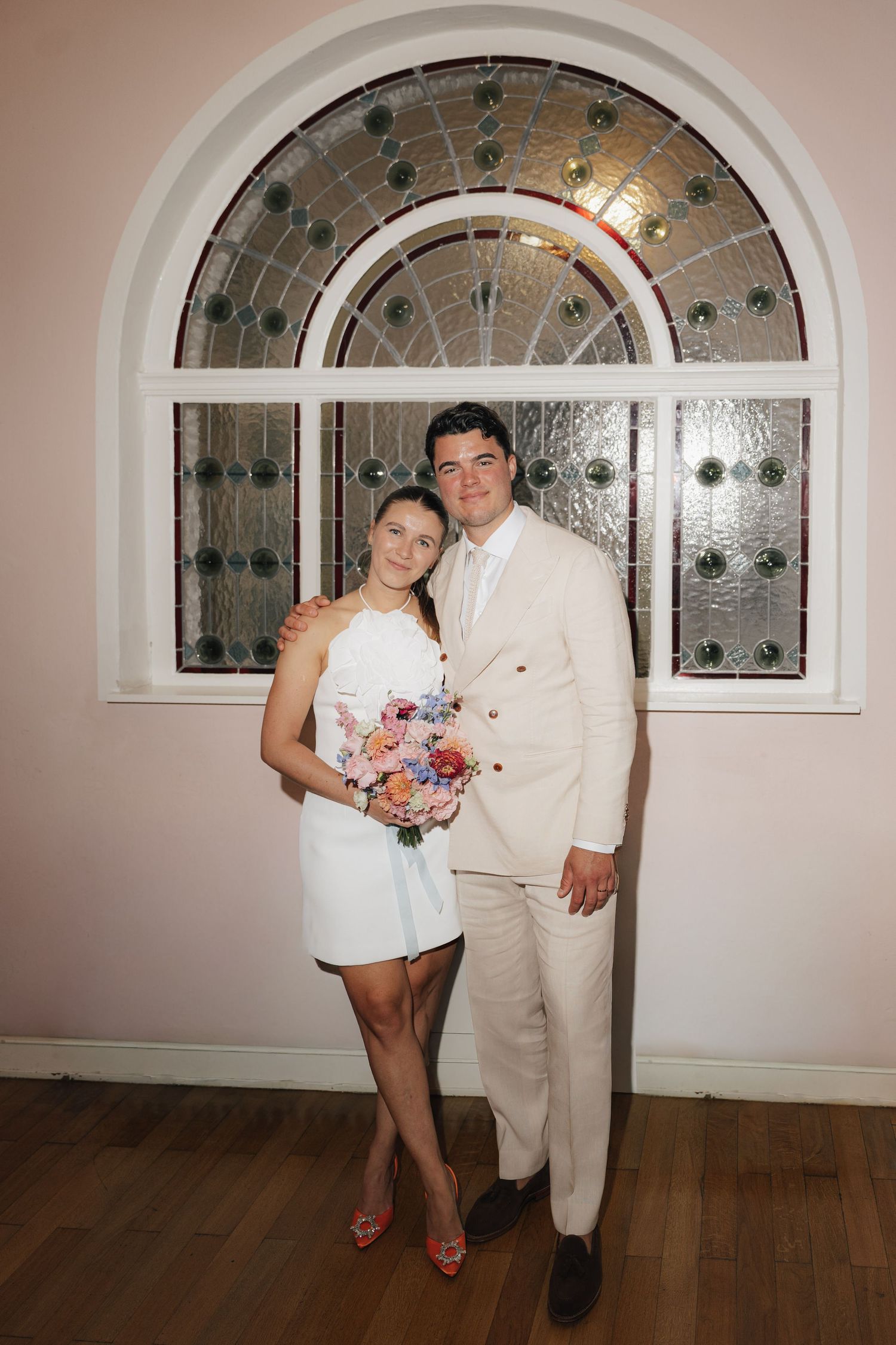 Couple posing in white attire against an arched window with decorative glass details.