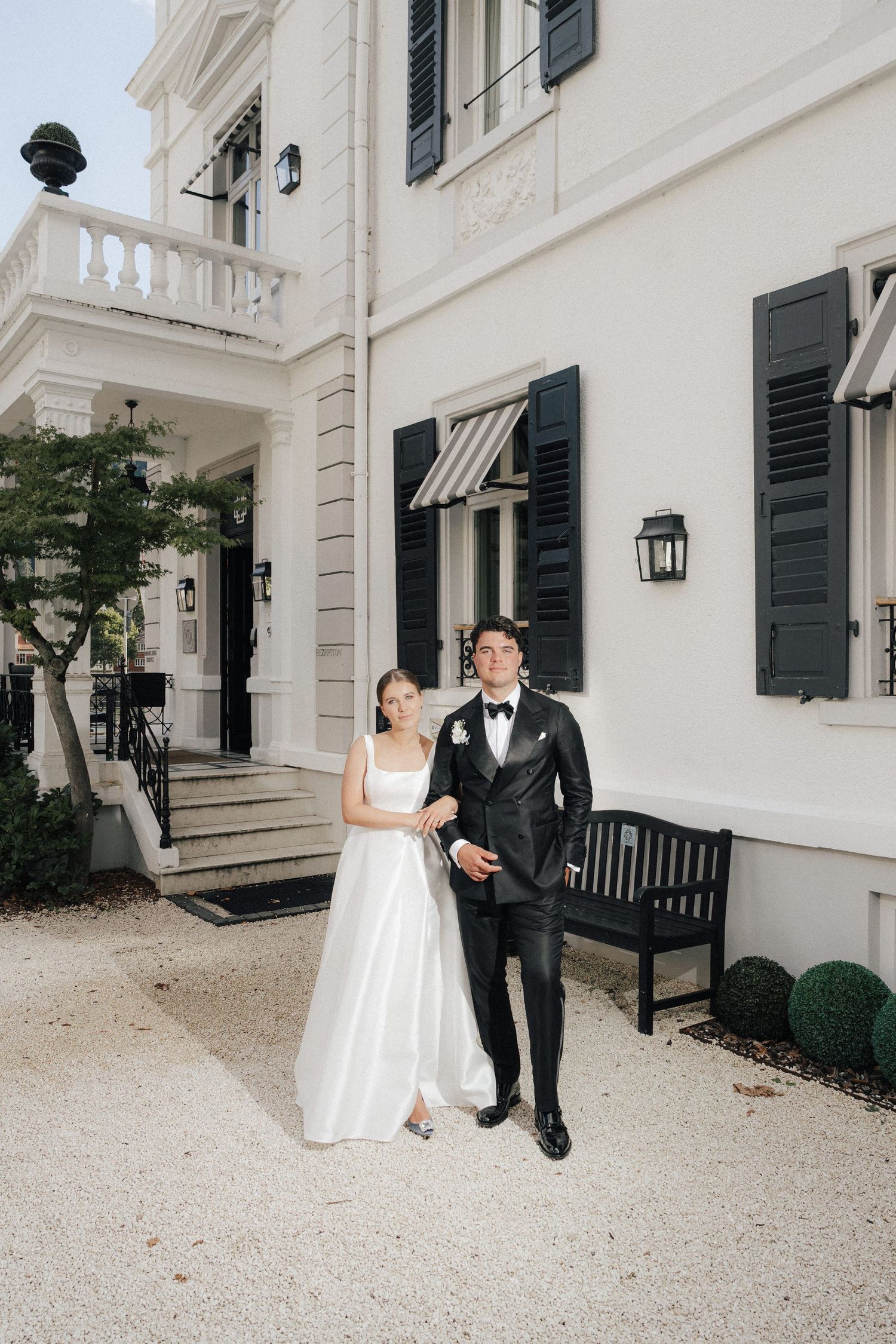 A wedding couple poses playfully against a white colonial style building with black shutters.