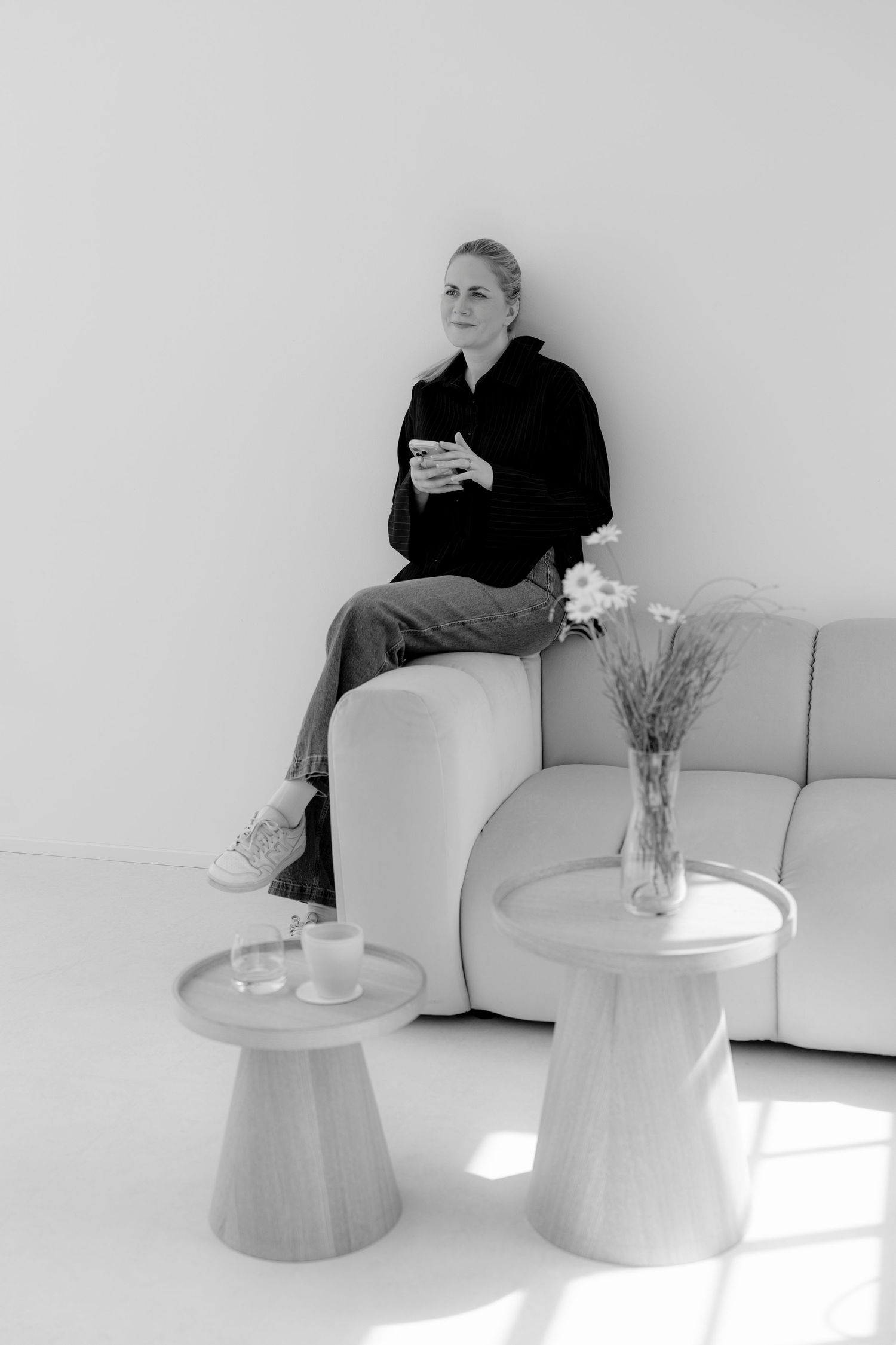 Black and white photo of a woman sitting on a modern sofa holding a phone, with small side tables and a vase nearby.