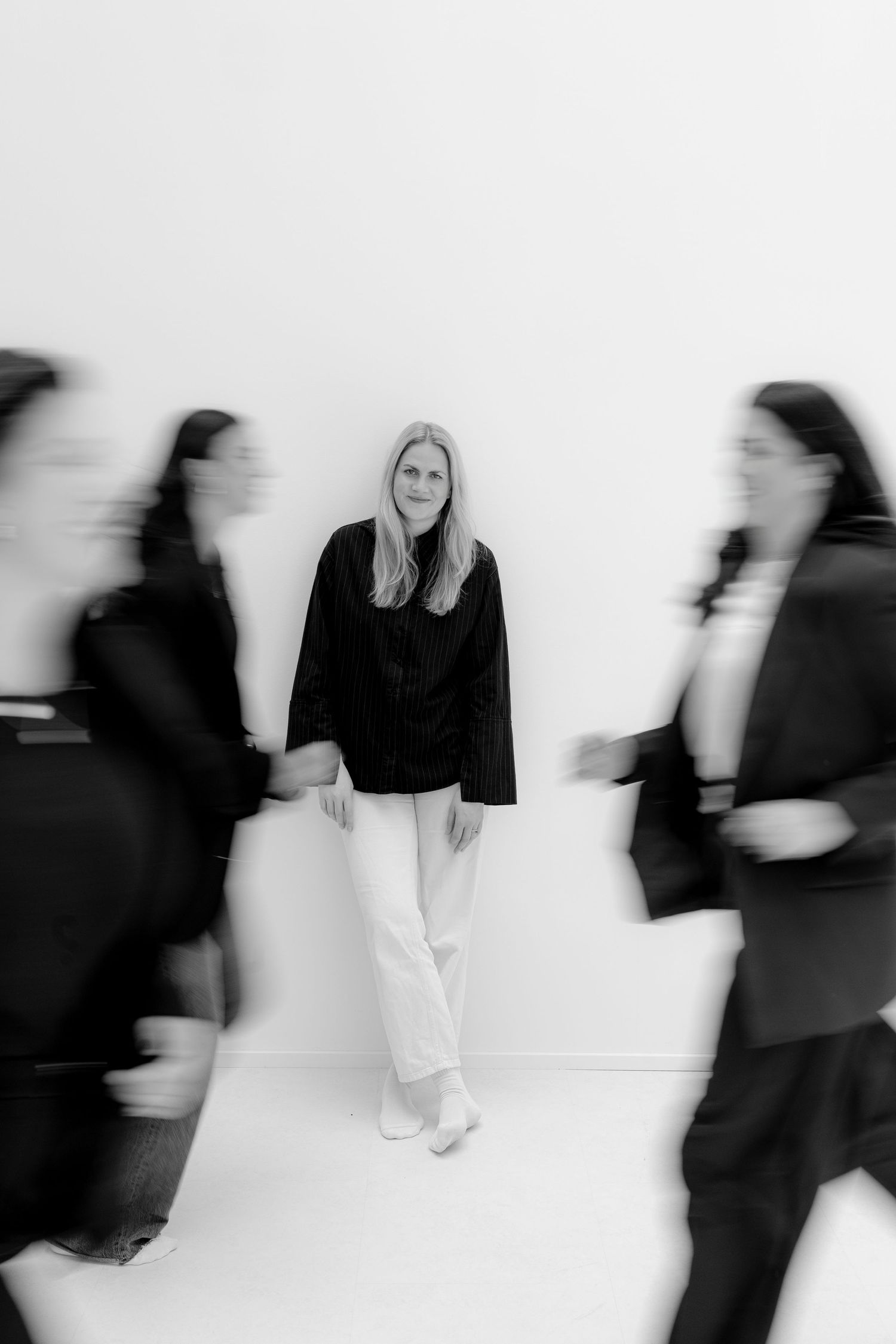 Black and white photo of a woman in white pants standing still against a wall while blurred figures in black rush past her.