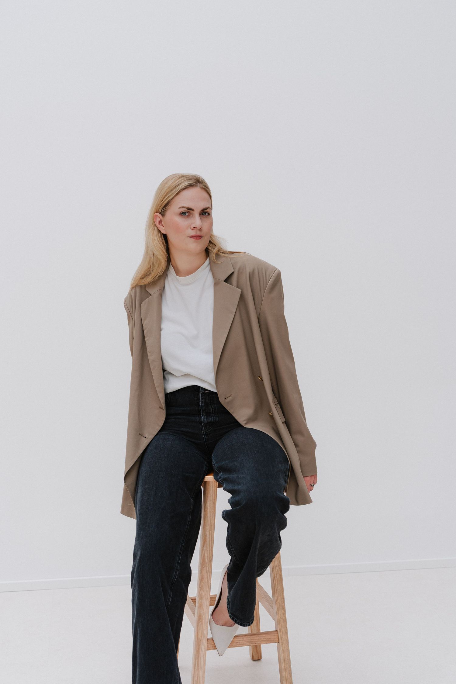 Woman in oversized beige blazer, white tee, and black jeans sitting on wooden stool against white background.
