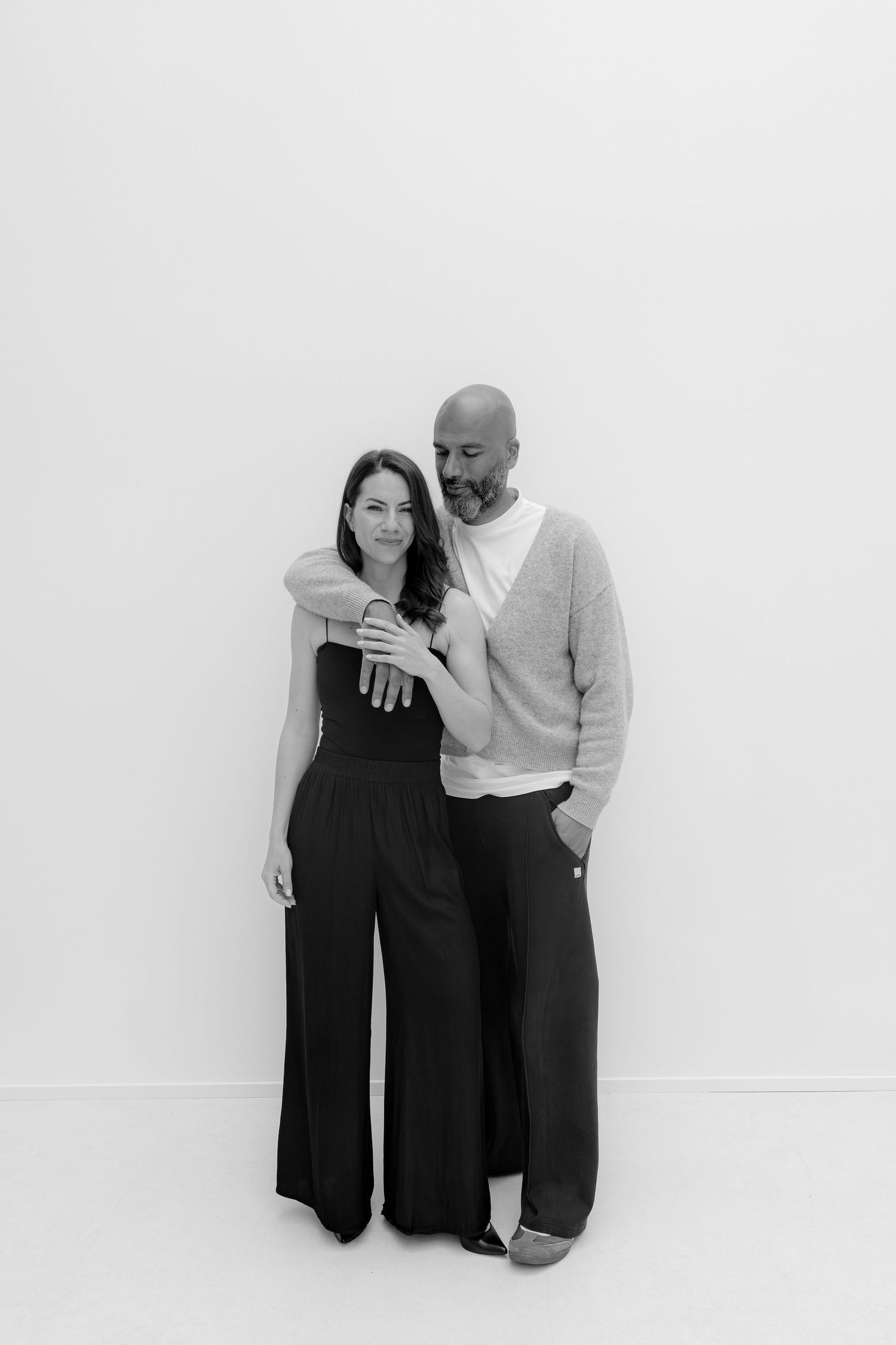Black and white portrait of a couple embracing, man in sweater with arm around woman in black jumpsuit.