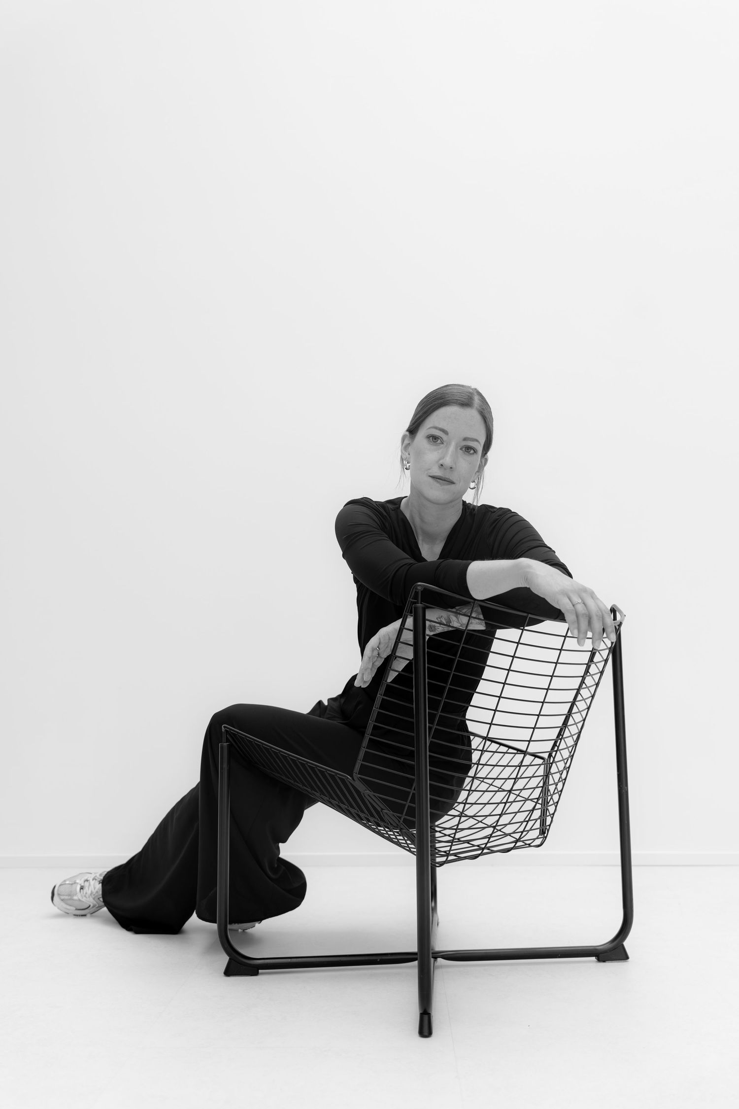Black and white photo of a woman in black outfit sitting relaxed on a wire chair against a white background.