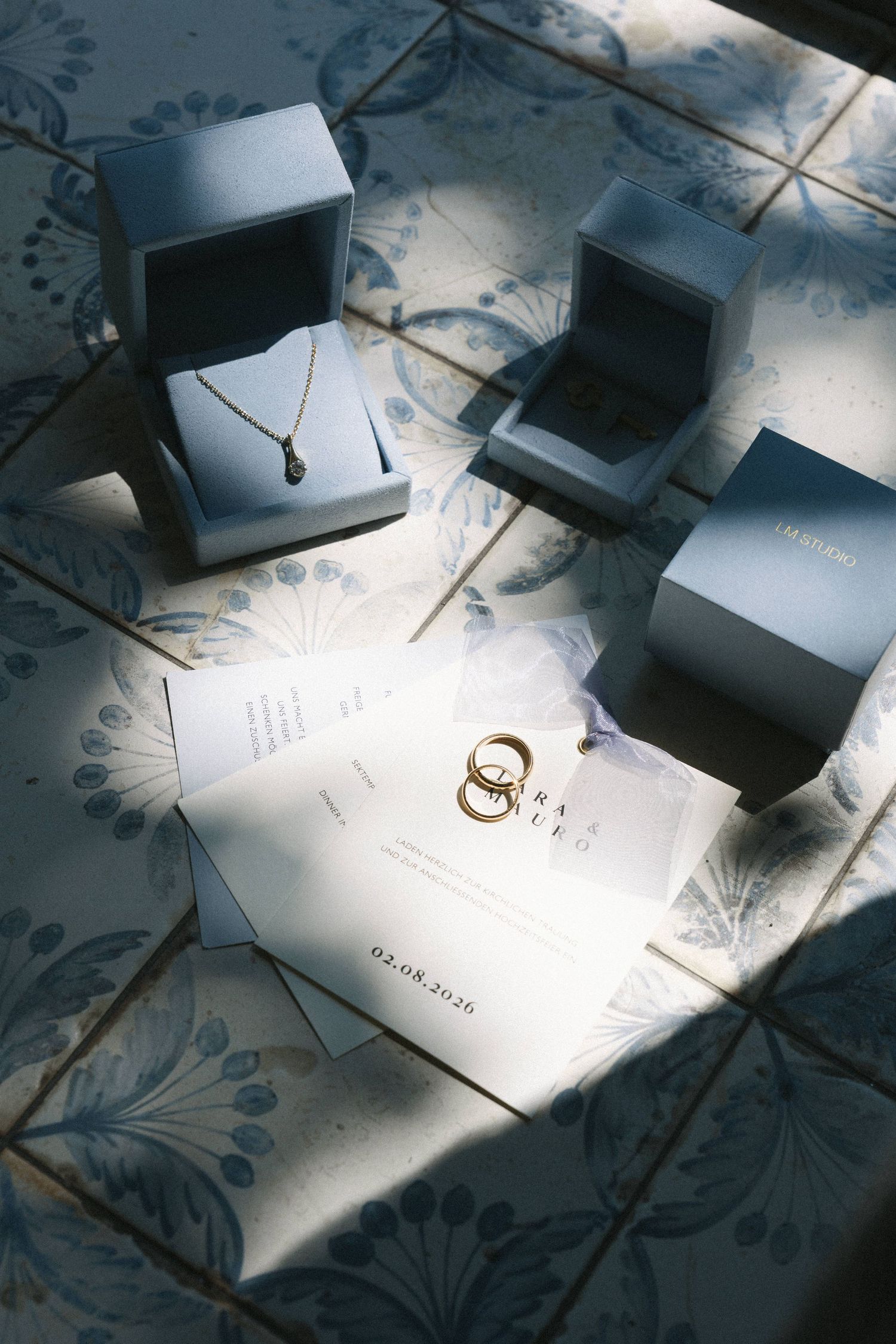 Elegant wedding jewelry display with gold necklace in blue box, wedding rings, and invitation on decorative tile.